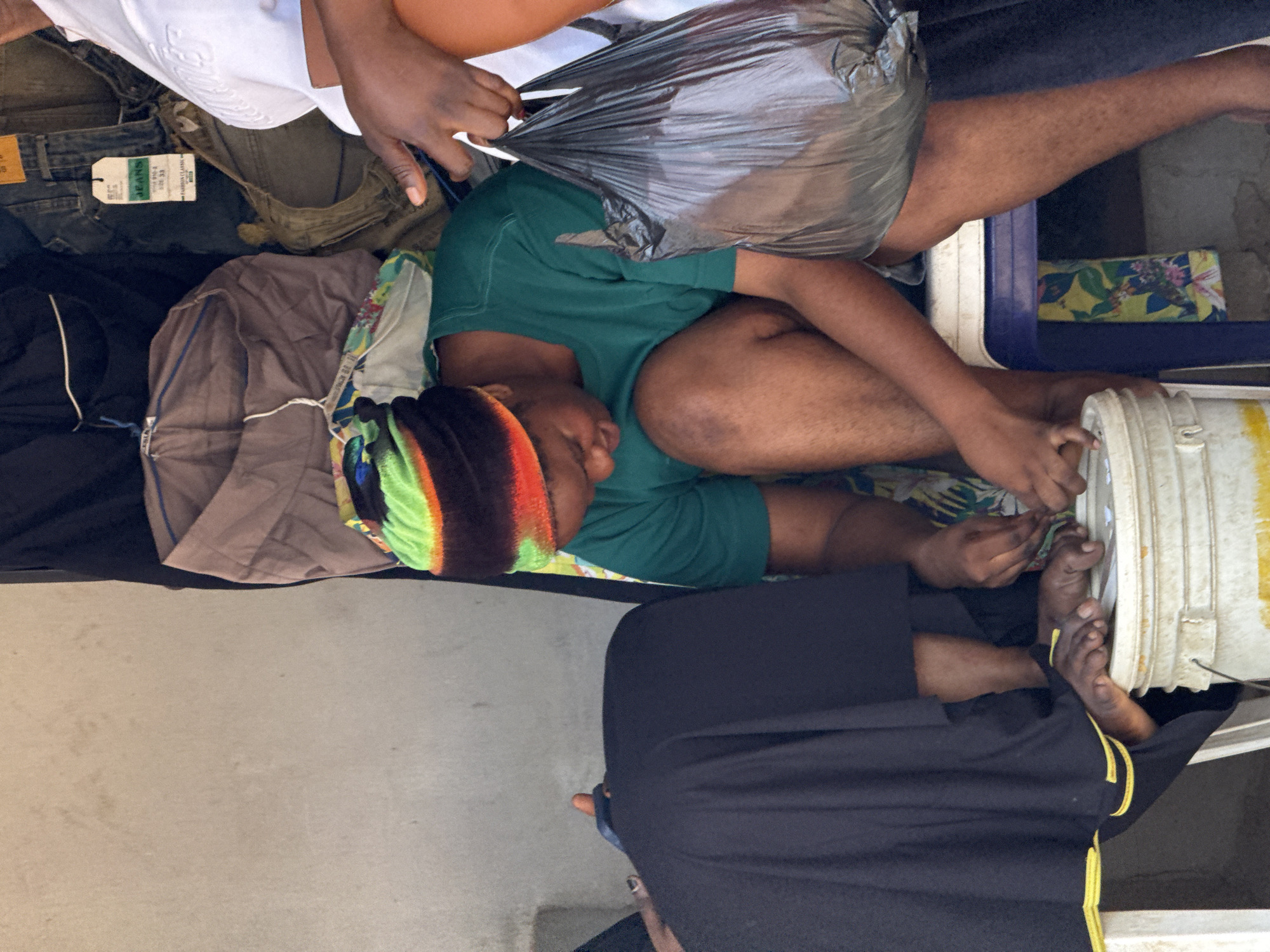 The image depicts an outdoor scene, likely a marketplace stall, in Abuja, Nigeria. In the foreground, a woman wearing a green t-shirt and a colorful knitted cap is seated, tending to her toenails. She is positioned behind a white bucket. To her left, another person, mostly obscured by dark fabric, appears to be seated or lying down. To the right, a person in a white shirt holds a black plastic bag, and their hands are visible near the woman with the green shirt, possibly interacting or handing something over. Behind them, a variety of clothing items, including jeans and folded fabrics, are displayed, suggesting a retail setting. The overall environment appears informal and somewhat cluttered.