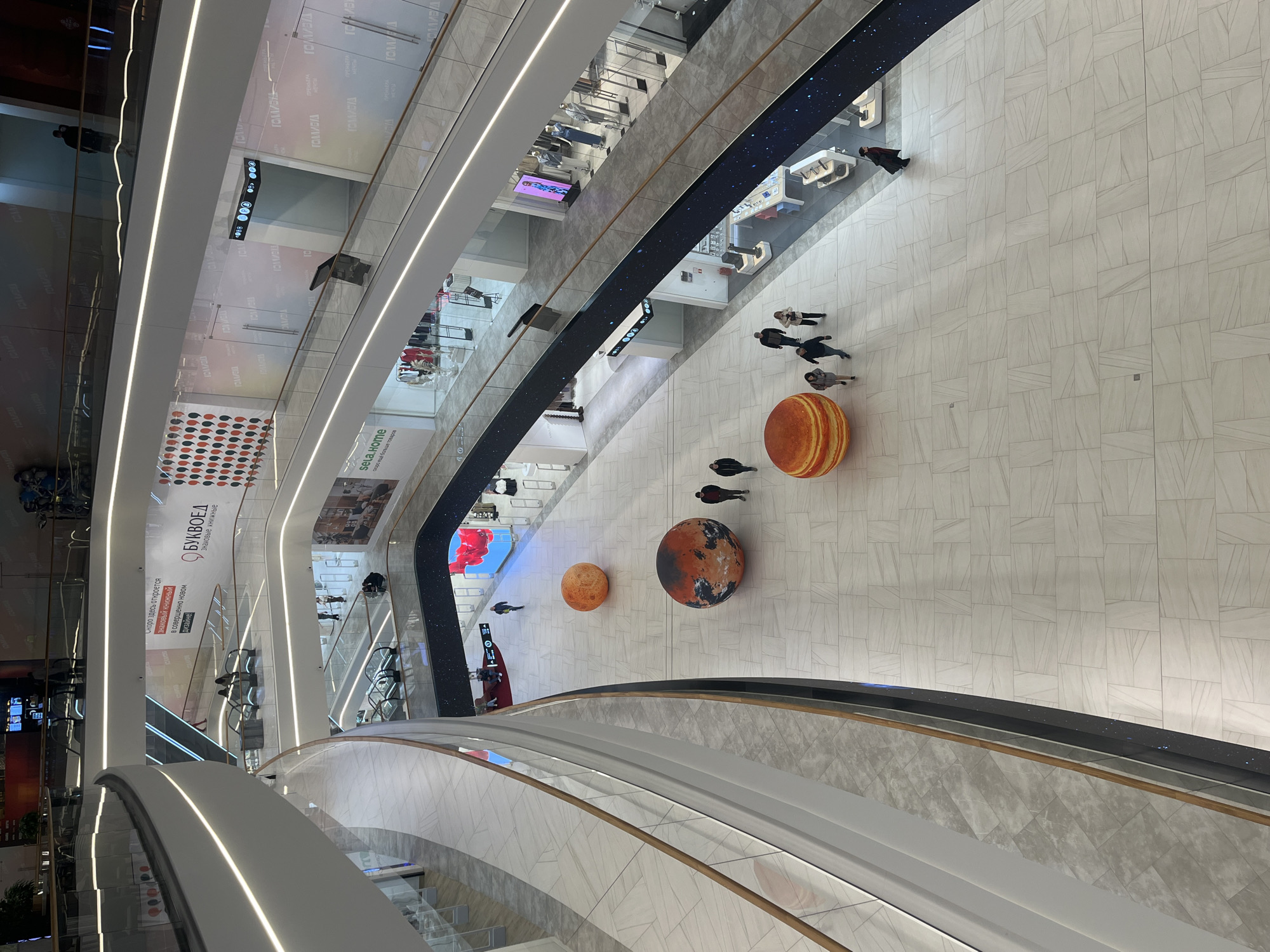 This is an indoor shot, likely taken from an elevated position within a multi-level shopping mall or retail space. The perspective is tilted and the photo is a high angle shot looking down.
Several floors of the mall are visible, interconnected by escalators. The flooring appears to be composed of light-colored, rectangular tiles. There are large, spherical objects on the floor, resembling stylized planets or celestial bodies, which are likely part of a decorative installation. People are walking around these objects, suggesting they are attractions or landmarks.
Along the edges of the floors are glass or transparent barriers, providing views into the retail spaces. Storefronts and branding are visible, including signs in Russian. There are also illuminated linear light fixtures and a dark strip with light points resembling stars. Escalators are visible in the foreground.