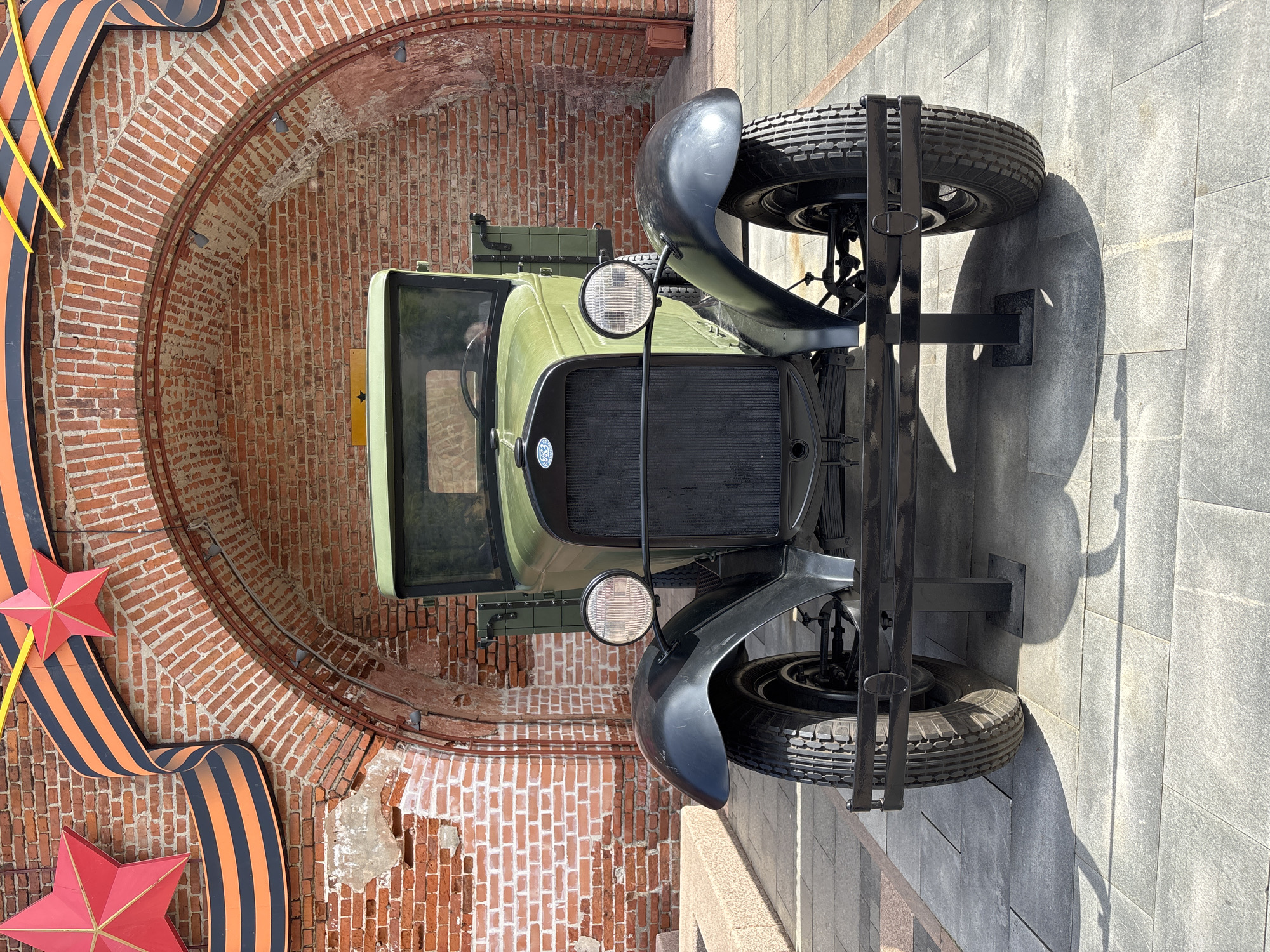 An antique olive-green truck, likely a Soviet-era model, is displayed prominently in a sunlit outdoor setting. The truck faces forward, showcasing its black grille, round headlights, and dark fenders. A spare tire is mounted vertically on a black stand to the right of the front bumper. The ground beneath and in front of the truck is paved with light grey, rectangular tiles, casting strong shadows from the vehicle and its stand, indicating bright daylight.
Behind the truck, a large brick archway or tunnel entrance forms the backdrop. The bricks are reddish-brown and show signs of age and some wear. To the left of the truck and archway, a large decorative display features a flowing black and orange striped ribbon, reminiscent of the St. George's ribbon, adorned with prominent red stars. These symbols are often associated with Victory Day celebrations in Russia, suggesting a commemorative or historical context for the display in Nizhny Novgorod.
The truck appears to be a static exhibit; no activity is taking place. A round emblem is visible on the center of the truck's grille, and a yellow sign with a black star is fixed to the brick wall behind the truck's cabin, though no text is clearly legible from this distance. The overall scene evokes a sense of historical remembrance in a public space.