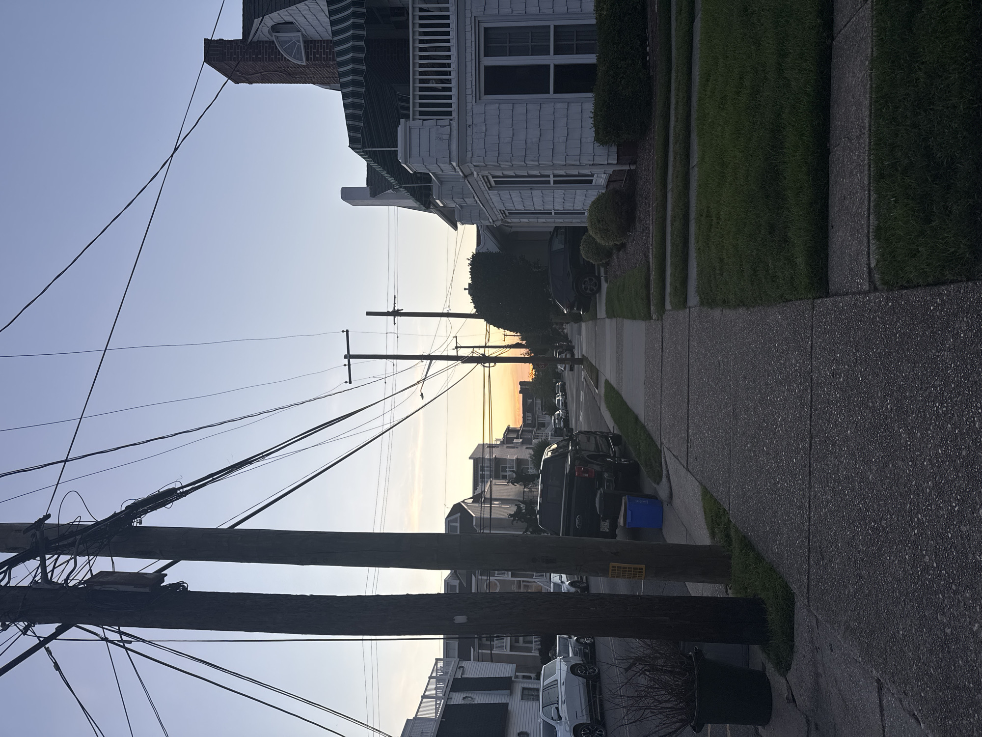 The image depicts a residential street in Ocean City, United States of America, during what appears to be sunset. The sky is clear with a soft gradient of orange and yellow near the horizon, transitioning to a pale blue overhead. A network of utility poles and wires dominates the foreground and mid-ground, casting shadows on the concrete sidewalk. On the right side of the sidewalk is a white, multi-story house with a green and white striped awning over the second-floor balcony. The house features shingled siding and several windows. Adjacent to the house is a well-maintained lawn with neatly trimmed bushes. On the left side of the sidewalk, across the street, are other residential buildings, some with balconies, and parked vehicles, including a white SUV and a dark-colored pickup truck. The street itself is paved, and the sidewalk is made of concrete. A blue recycling bin is visible near the curb next to the pickup truck. The overall scene suggests a quiet, suburban neighborhood at dusk.