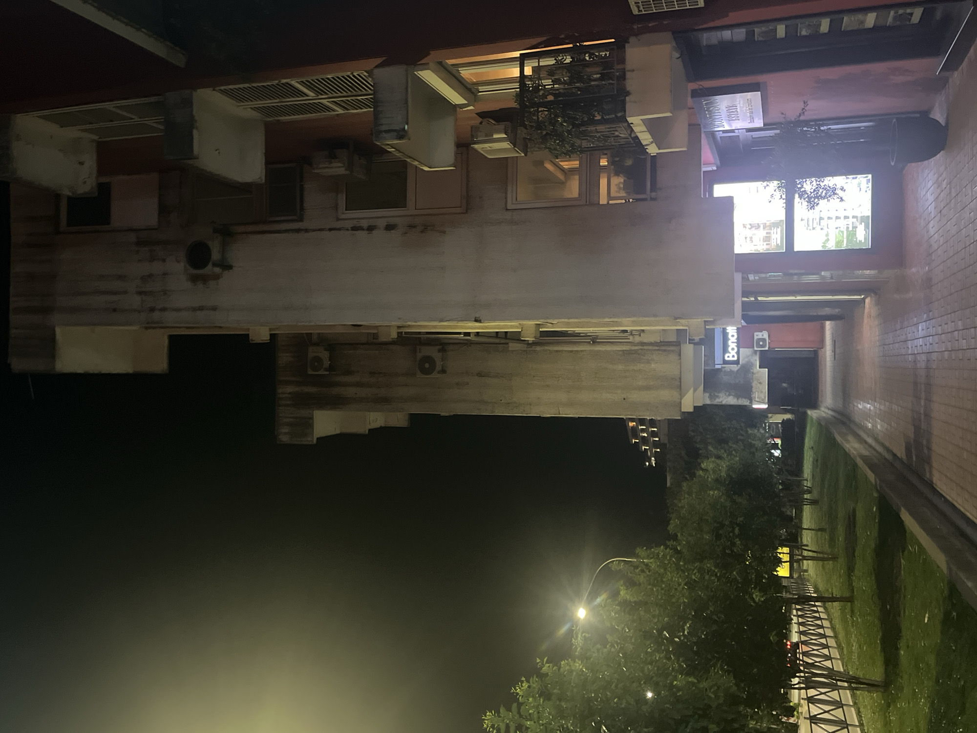 A nighttime urban scene in Budva, Montenegro, reveals a narrow passage between buildings. The light from a street lamp illuminates the trees and grass. Buildings on either side exhibit residential architecture, with windows, balconies, and air conditioning units. A sign bearing the name "Bonatt" is visible on one building. Paved pedestrian walkways are adjacent to the grassy area with trees.
