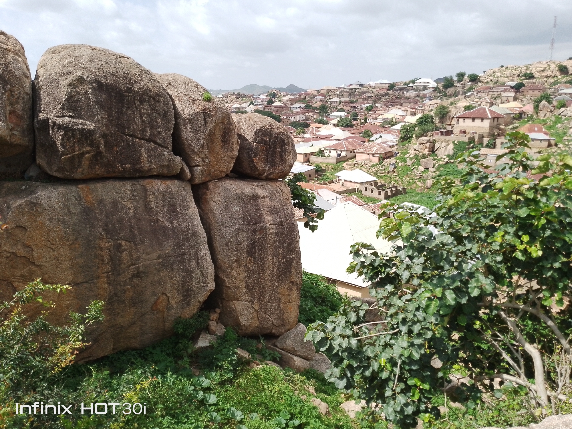 This is a medium shot, exterior, daytime, eye-level view of a rocky landscape with a village in the background. In the foreground, a cluster of large, weathered boulders dominates the left side of the frame. These boulders are textured and vary in color from dark brown to lighter grey, with some patches of moss or lichen. To the right of the boulders, a lush green tree with broad leaves partially obscures the view.
Beyond the boulders and the tree, a densely populated village spreads across a hilly terrain. The village is characterized by numerous buildings with corrugated metal roofs in shades of orange and grey, interspersed with lighter-colored walls. The houses are clustered together and appear to be built on and around the rocky landscape. The terrain is uneven, with exposed rocks and patches of green vegetation visible throughout the settlement. In the distance, rolling hills can be seen under a cloudy sky. The overall impression is of a rural settlement integrated with its natural rocky environment. A watermark at the bottom left of the image reads "Infinix HOT 30i".
