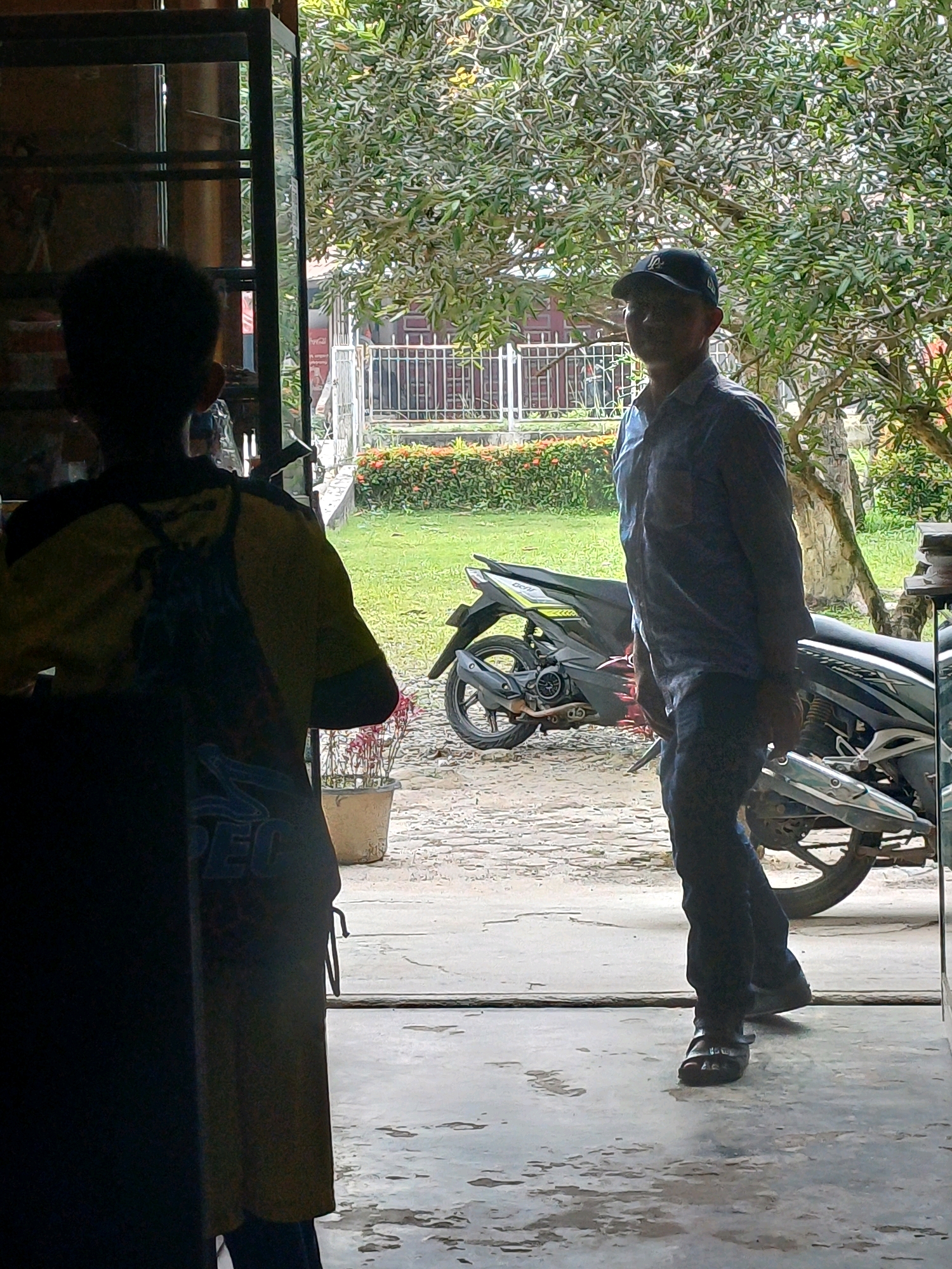 A man wearing a dark baseball cap and a light blue collared shirt, dark pants, and sandals stands on a concrete walkway. He is facing slightly to his left, looking towards the viewer. Behind him, partially obscured by a tree with green foliage, is a fence and a building. To his right, two motorcycles are parked on the walkway. In the foreground, to the left, a person wearing a yellow and black shirt and a backpack is silhouetted and facing away from the viewer. The scene appears to be outdoors, possibly in a residential or commercial area of Dumai City, Indonesia.