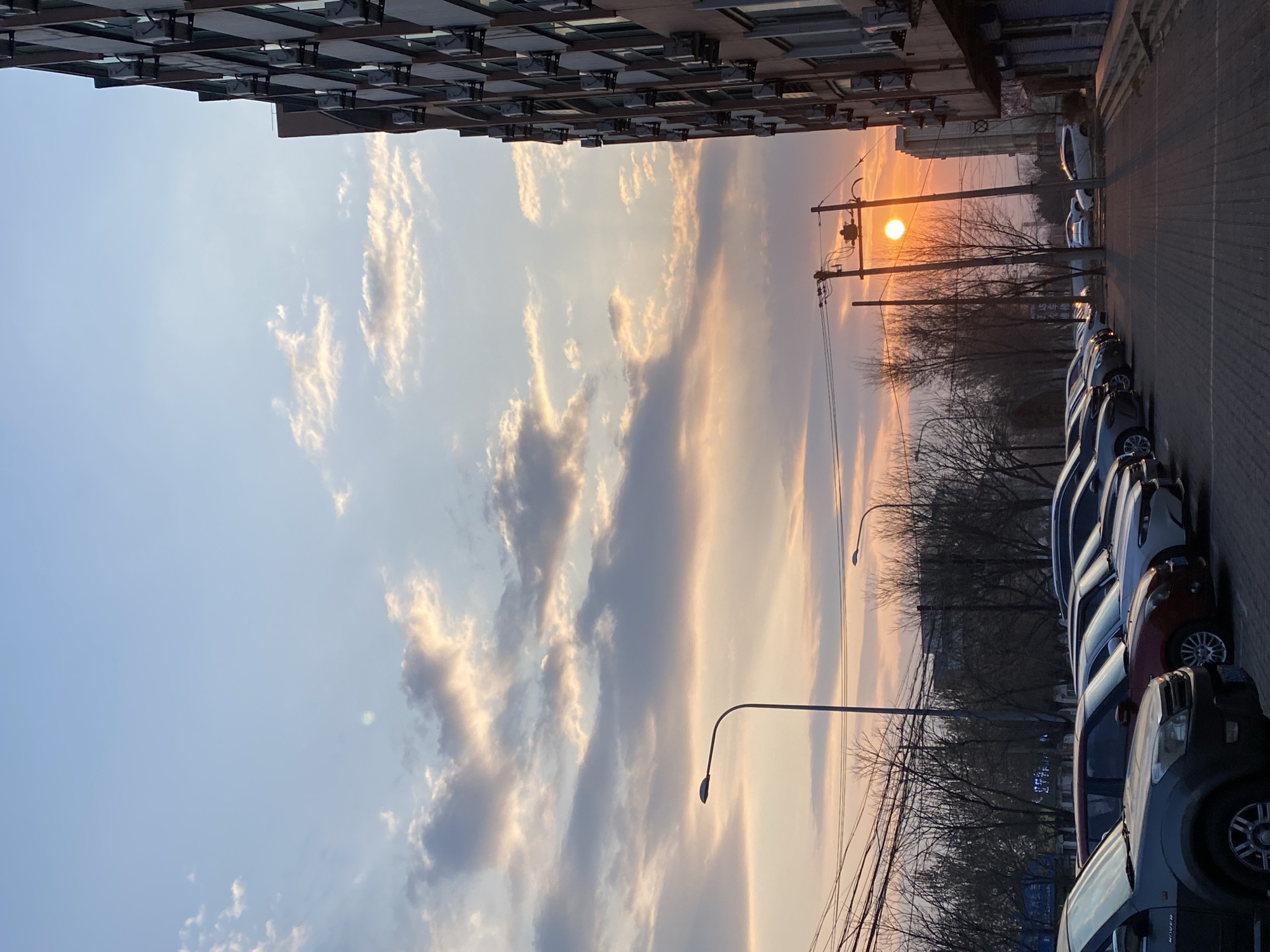 The image shows a street scene in Zhongtun, China at sunset. A tall building is on the left, and cars are parked along the street on the right. Telephone poles and bare trees are visible in the middle ground, with the sun setting in the distance, creating an orange glow in the sky. The sky itself is filled with clouds.