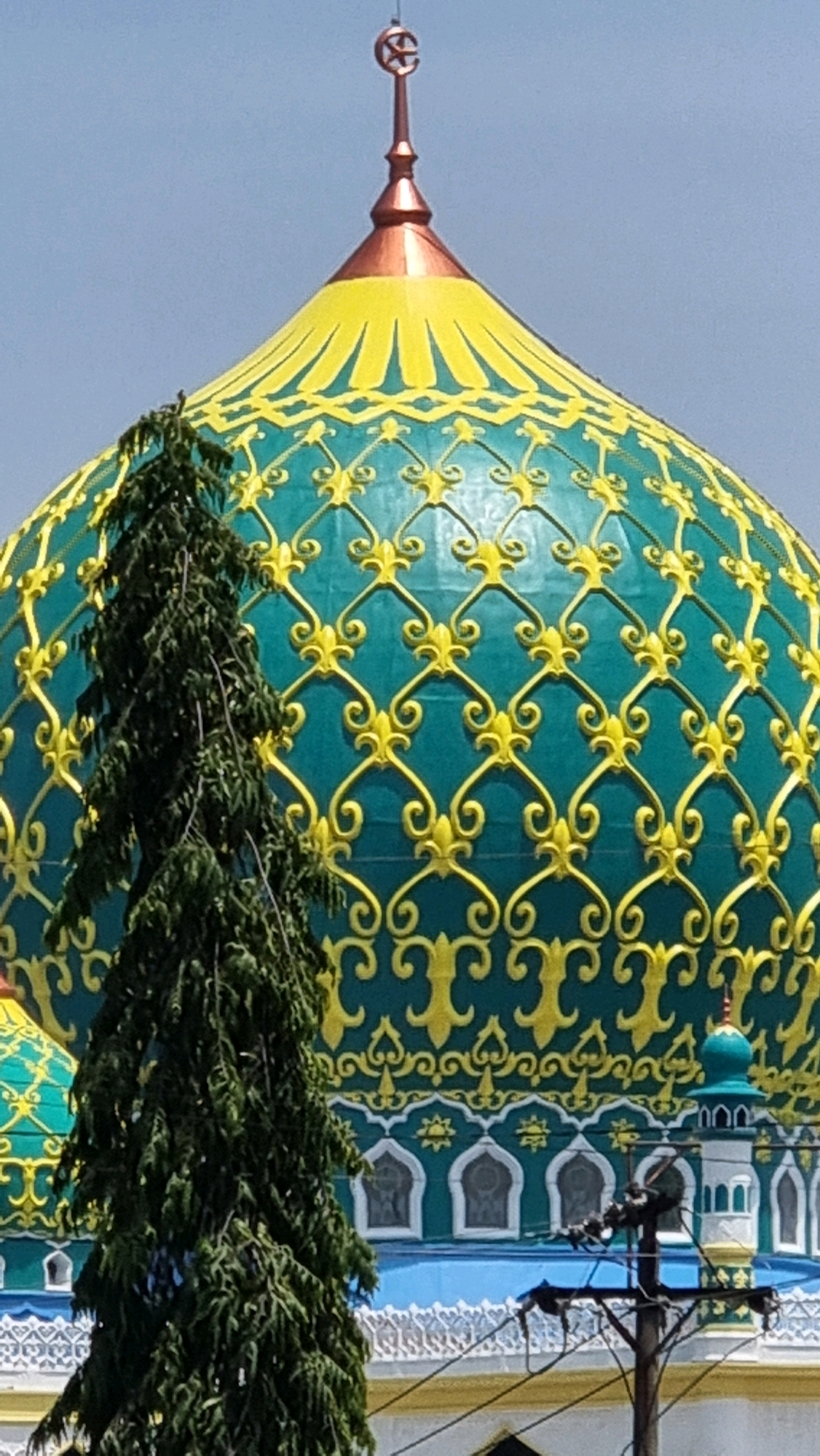 The image captures a close-up view of the turquoise and gold dome of a mosque, likely located in Lhokseumawe, Indonesia. The dome is adorned with intricate yellow patterns that form a repeating grid of floral and swirling motifs. At the apex of the dome is a copper-colored finial topped with a crescent moon symbol. To the left, a tall, slender evergreen tree partially obscures the view of the dome. Below the main dome, a white structure with arched windows and a smaller turquoise dome with a white minaret is visible, suggesting the presence of a larger mosque complex. Electrical wires and a utility pole are also present in the foreground. The sky is a clear, light blue, indicating a sunny day.