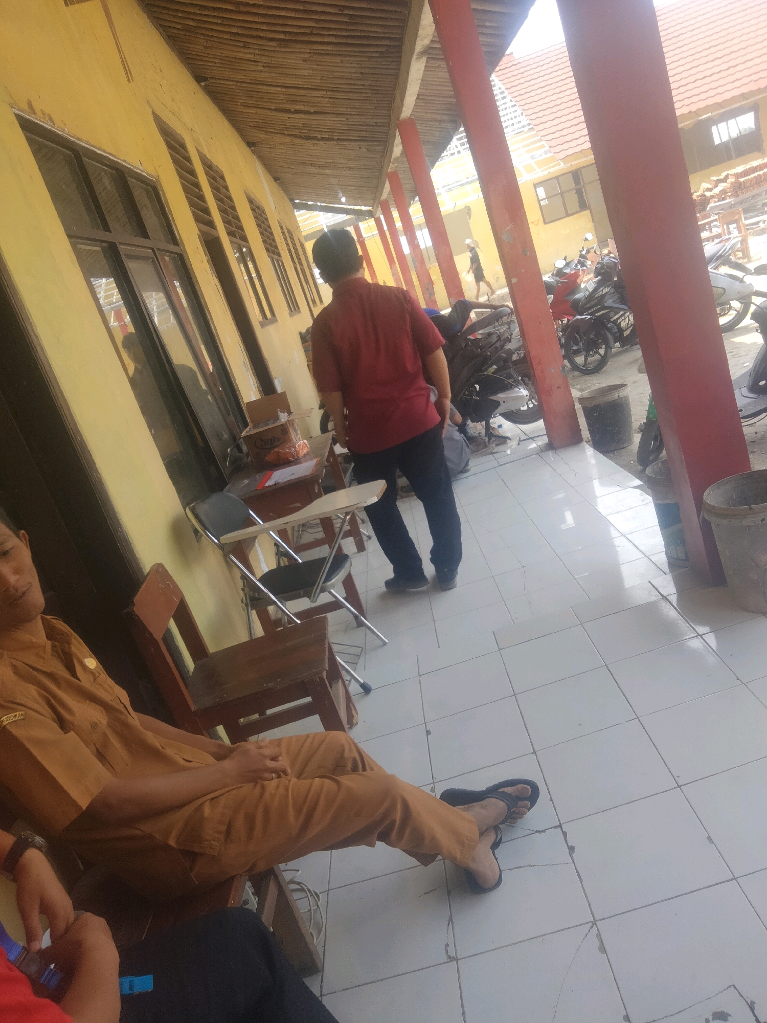 The image depicts an outdoor scene in Suka Sari, Indonesia. In the foreground, a person wearing a tan uniform is seated on a wooden chair. The uniform appears to be a standard Indonesian civil servant attire, characterized by its brown color and insignia on the left chest pocket. The person's legs are crossed, and they are wearing black flip-flops. To the left of this seated individual, another person's hands are visible, suggesting they are interacting with something.
In the middle ground, a man in a red short-sleeved shirt and dark pants is standing and facing away from the camera. He is positioned near a desk with a box on it. Several foldable chairs, some with attached writing surfaces, are also present. Beyond the man, a row of motorcycles is parked. The environment includes a building with a yellow facade and wooden-slatted eaves. Large red pillars support an overhang, and the ground is tiled with white squares. In the background, more motorcycles are visible, along with glimpses of other buildings and what appears to be roof tiles. The overall scene suggests a public or semi-public area, possibly a school or government office, given the uniforms and the presence of desks and chairs.