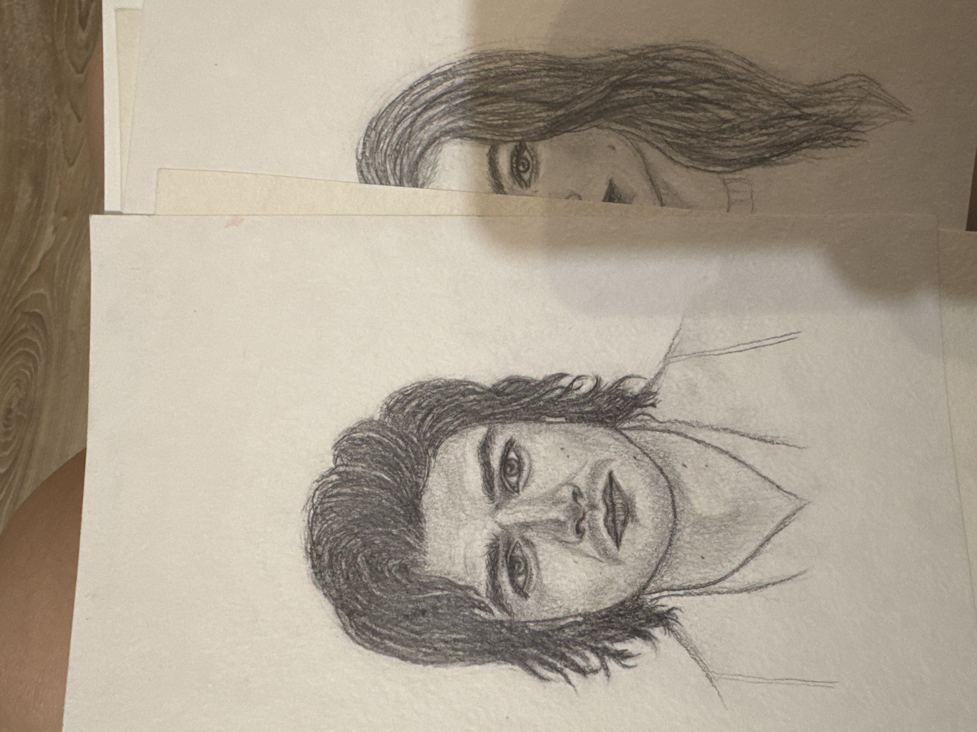 Two graphite pencil portraits are displayed on a light-colored paper. The portrait on the left depicts a young man's face and upper chest. He has dark, wavy hair, prominent eyebrows, and a serious expression. The portrait on the right shows the right side of a young woman's face and hair. She has dark, long hair and her eye is clearly visible. The two drawings are placed side-by-side, with the woman's portrait partially overlapping the man's. The background appears to be a wooden surface. There is no indication of the location being Mikhaylovsk, Russia.