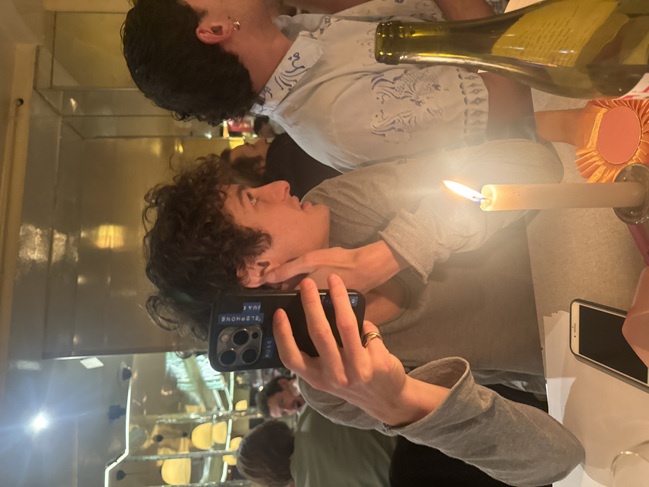 A person with curly hair takes a selfie in a mirror with a phone, while another person in a patterned shirt appears above in the reflection. A lit candle stands between them, casting light. A wine bottle is also visible near the top of the frame. A second phone lies on the table to the right. The restaurant setting is discernible in the mirror's background.