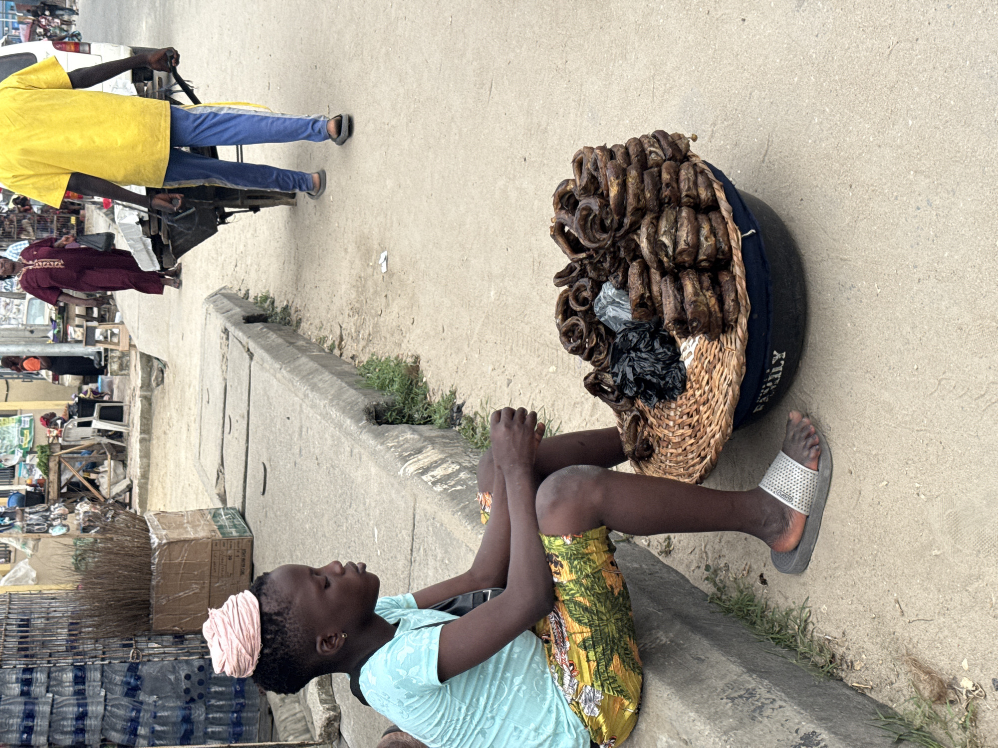 In Ado, Nigeria, a young Black girl with a pink headwrap sits on a concrete curb next to a basket of dried fish. She wears a light blue t-shirt and colorful patterned shorts. Her gaze is directed downwards, and her hands are clasped in her lap. To her right, a large woven basket overflows with stacks of circular, dried fish, some wrapped in black plastic. The ground is paved with concrete, and a concrete drainage channel runs along the curb. In the background, a stall displays bottles of water, bundles of dried reeds, and various goods. A person in a yellow shirt pushes a cart, and other individuals are visible walking in the distance. The scene depicts a street market environment.