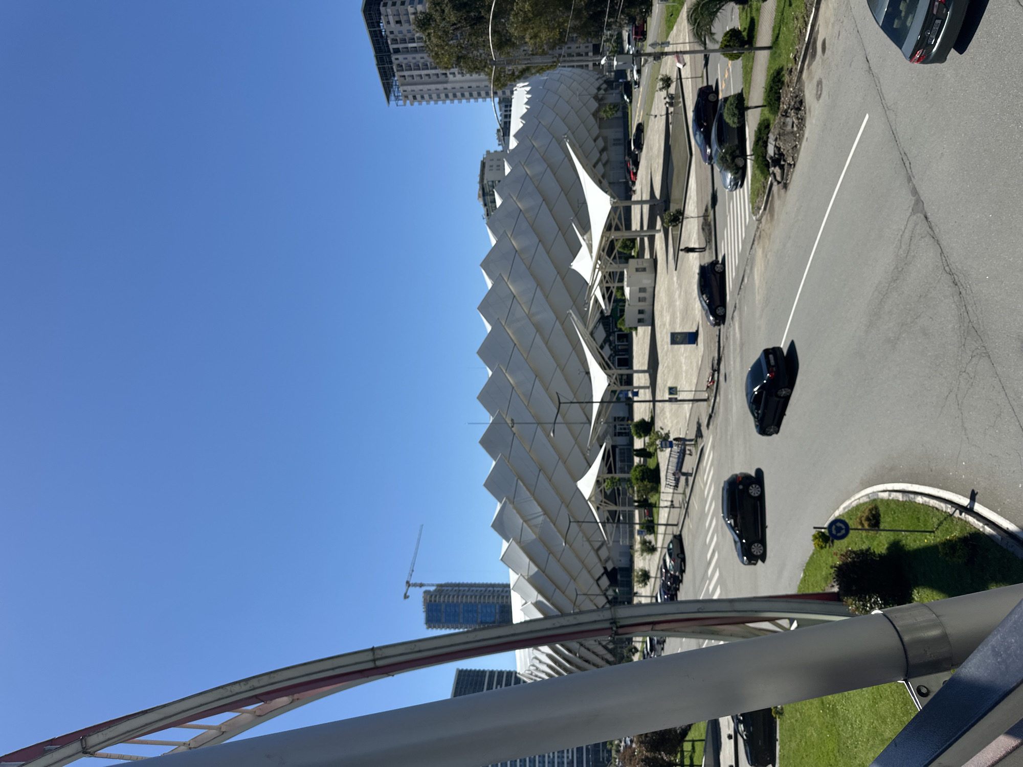 The image shows a view of Batumi, Georgia, with a distinctive white, geometrically patterned structure dominating the scene. The structure is accompanied by a series of white tensile canopies supported by metal frames. A road with parked cars and crosswalks runs alongside this structure. In the background, several high-rise buildings are visible, including one under construction with a crane. A clear blue sky occupies much of the upper part of the image. The shot appears to be taken from an elevated position, possibly a bridge, with part of the bridge's structure visible in the foreground. The composition and perspective suggest an urban setting.