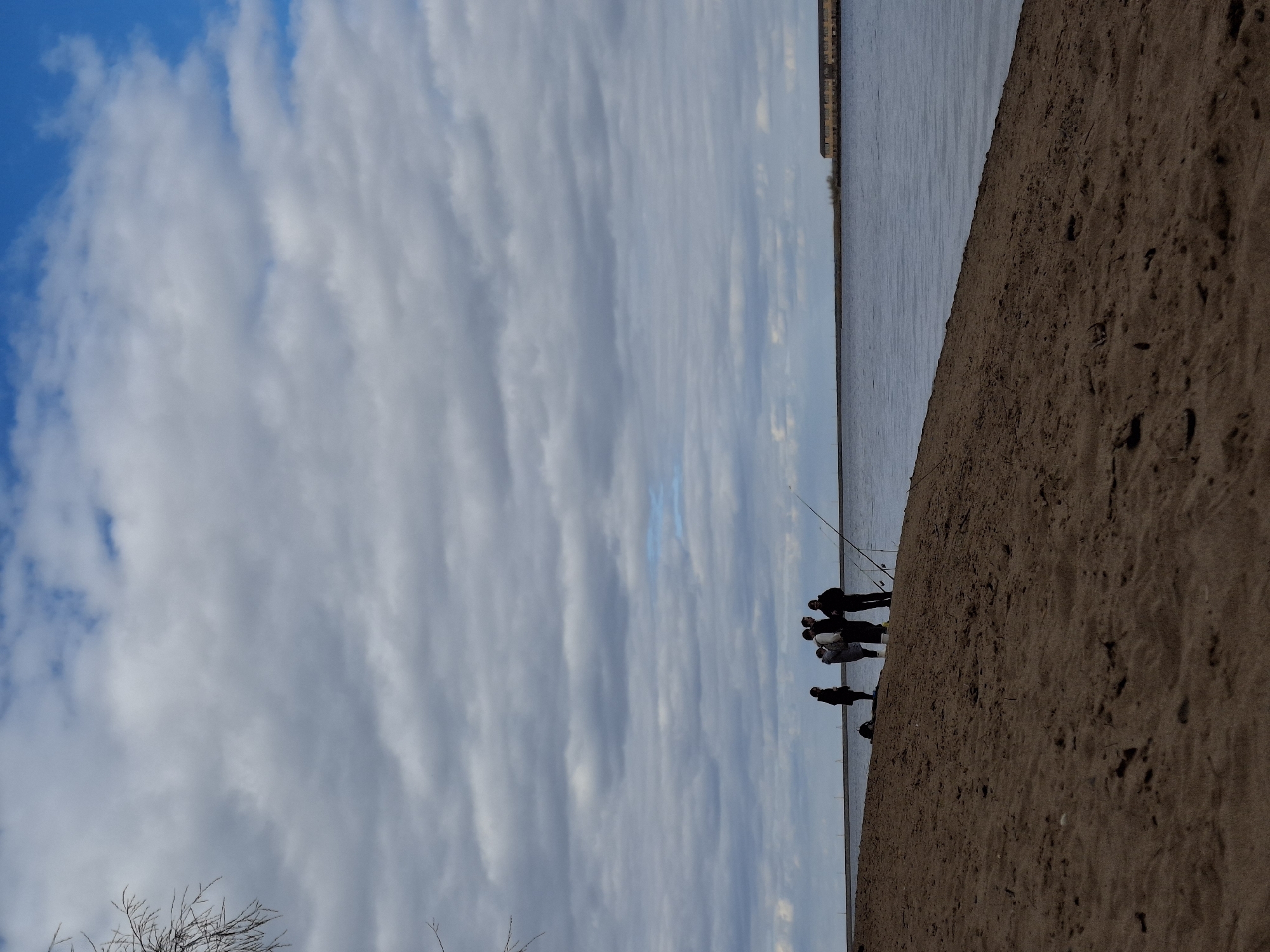 The image shows a group of people fishing by a body of water. They are standing on a sandy bank. The sky is partly cloudy with patches of blue. The location is most likely Chaikovsky, Russia, based on the prompt. There are at least six individuals visible, and fishing rods are set up near the water's edge. The environment consists of sand, water, and clouds. The people are interacting by fishing together.