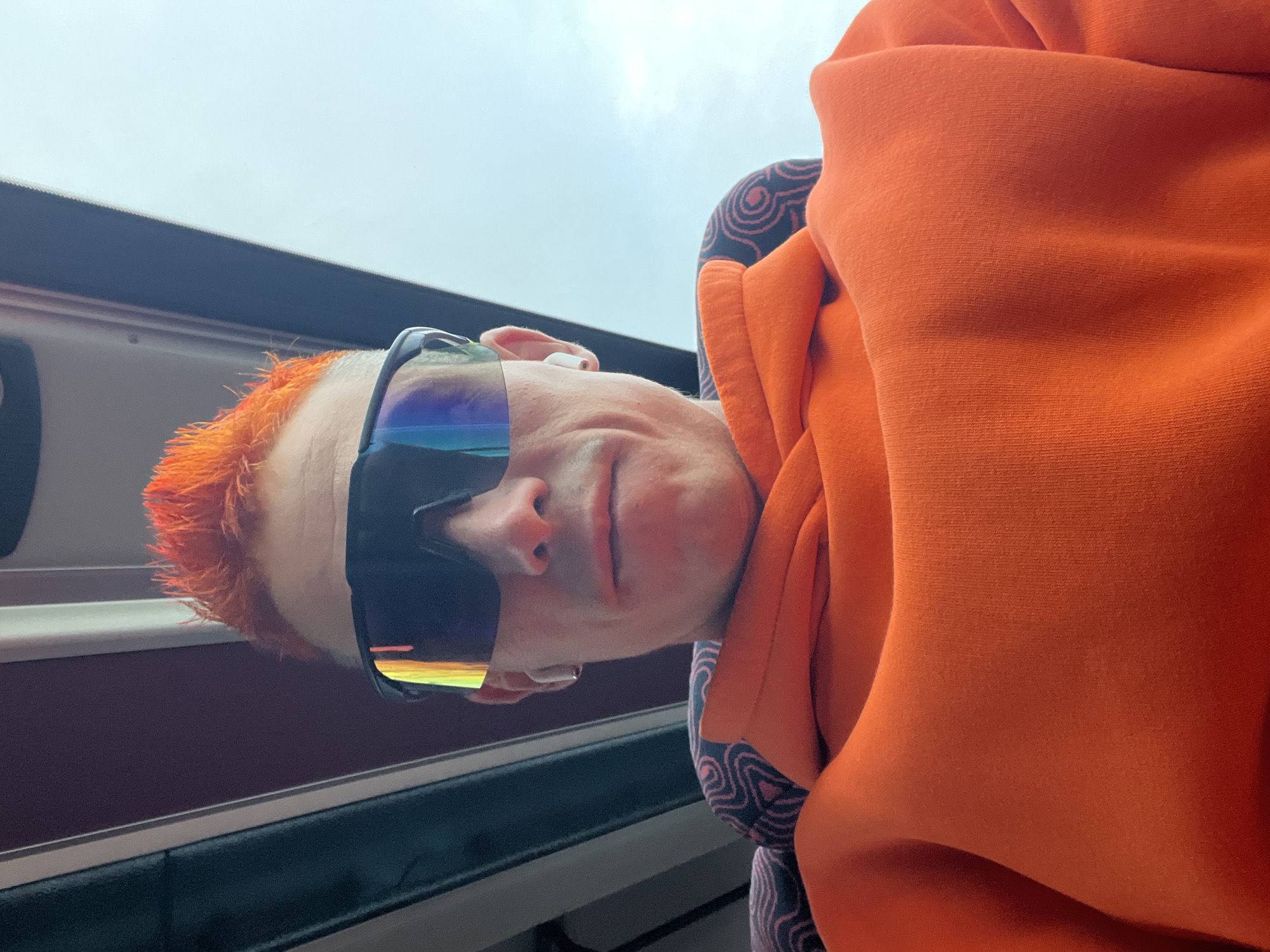 A close-up, low-angle selfie shows a person with bright orange, spiked hair wearing large, reflective sunglasses and a white earbud. The person is also wearing an orange hooded sweatshirt, the hood of which is pulled up and bunched around their neck. The background appears to be the interior of a bus or train, with dark seats visible on either side of the person and a bright, overcast sky visible through a window above. The image is taken indoors, and there is no indication of the city of Chaikovsky, Russia.