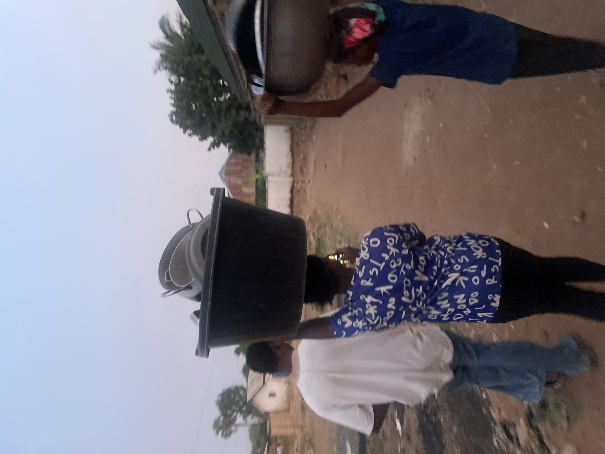 The image depicts three people walking on a dirt path outdoors. One person is wearing a white shirt and blue jeans. A second person is wearing a blue and white patterned shirt and black pants, carrying a stack of black buckets on their head. The third person is wearing a blue shirt and black pants, carrying a large metal bowl or pot on their head. The location may be in Jos, Nigeria; however, this is not verifiable from the image alone. The sky is overcast. Buildings and trees are visible in the background.
