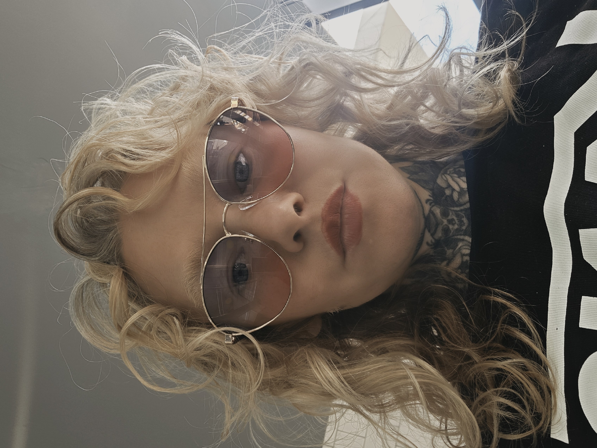 The image is a close-up, frontal selfie of a woman with blonde, curly hair. She is wearing large aviator-style sunglasses with rose-tinted lenses and a black t-shirt with a white graphic. She has visible tattoos on her neck and chest area. The lighting suggests an indoor setting, possibly a car, with diffused light coming from the right side of the frame. There is no indication of the specific location within Vsevolozhsk, Russia, or any specific action or interaction occurring. The environment appears to be plain and nondescript.
