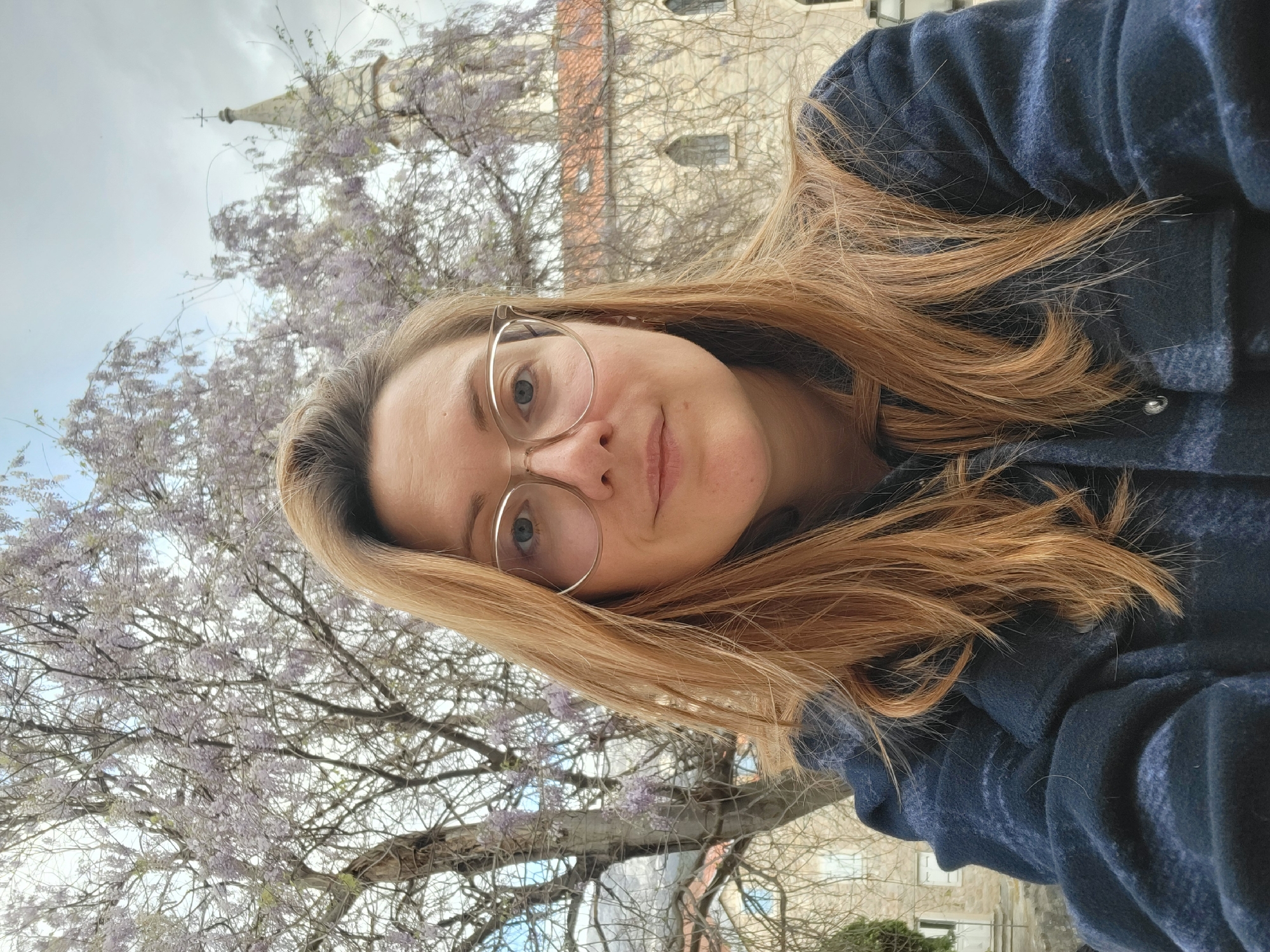 A fair-skinned woman with blonde/light brown hair, wearing eyeglasses and a blue patterned jacket, is taking a selfie outdoors. In the background is a flowering tree, possibly wisteria, and a stone building, likely a church tower. Given the location in Budva, Montenegro, the stone building is most likely the Church of St. Ivan, part of the Old Town architecture. The sky is overcast.