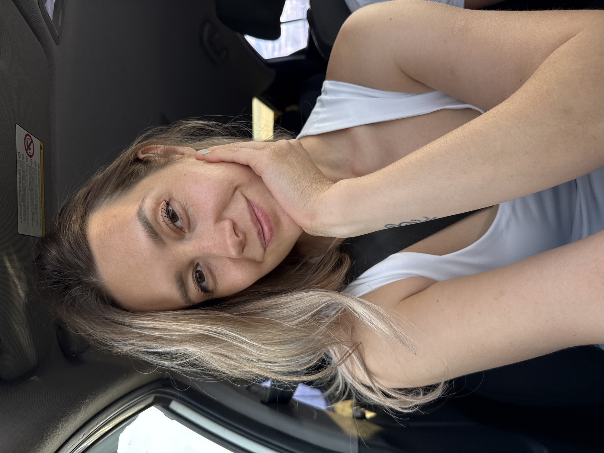 A young woman is taking a selfie in the passenger seat of a car. She has blonde hair and is wearing a white tank top. Her left hand is resting on her cheek, and she is smiling at the camera. The car appears to be parked, as the background is blurred. It is not possible to determine the specific location within Ust-Synya, Russia based on the image.