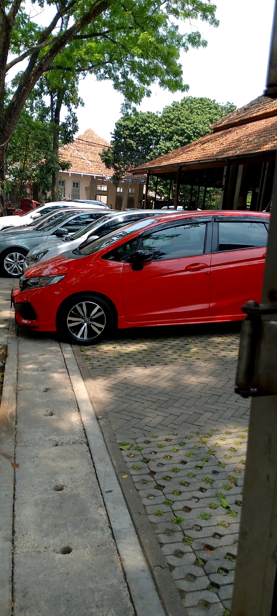 This is a medium shot, eye-level, outdoor photograph of a row of parked cars. The primary subject is a bright red hatchback in the foreground, positioned on the right side of the frame. Behind it, in a receding line, are several other cars, mostly silver or white. The cars are parked on a paved area composed of interlocking concrete blocks with grass growing in the gaps. To the left of the parking area is a concrete curb and drainage channel. In the background, a building with a tiled, brown roof and visible pillars is present, suggesting a covered outdoor area or veranda. Trees with lush green foliage are visible above and behind the building, casting shadows on the scene. The sky is bright and appears to be clear, indicating daylight. The overall environment suggests a parking lot adjacent to a building, possibly a restaurant, cafe, or resort, located in a tropical or subtropical climate. The presence of a red car, silver cars, a tiled roof building, and abundant green trees are consistent with a location in Bandung City, Indonesia. There are no people clearly visible in the frame, only the cars and the environment.