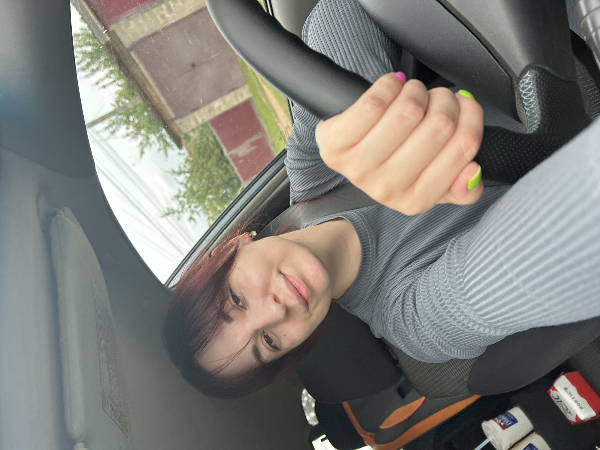 A young woman with auburn hair and bangs is sitting in the driver's seat of a car. She is wearing a gray, ribbed, long-sleeved shirt. Her left hand is on the steering wheel, and her fingernails are painted bright neon green. Her right hand is resting on the steering wheel, with fingernails painted neon pink. She is smiling at the camera. The interior of the car is visible, including the steering wheel, dashboard, and car seats. Outside the car window, a brick garage and trees are visible. The location is identified as Fokino, Russia.