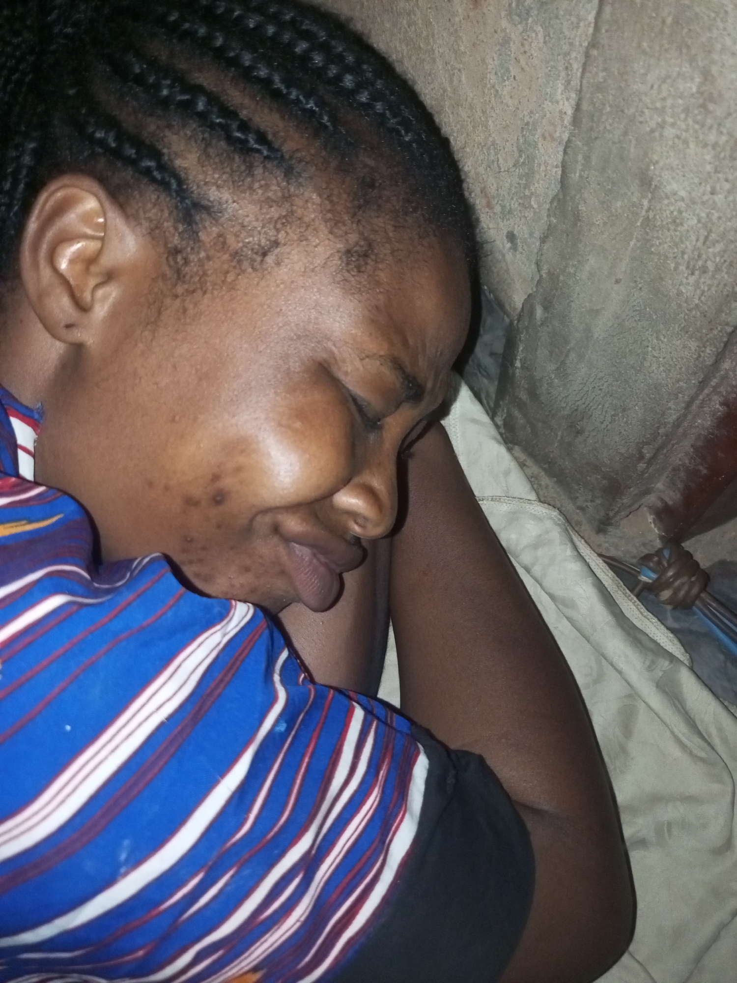 A close-up, overhead shot shows the side profile of a young Black woman sleeping. She is lying on her right side with her head resting on her right arm. Her eyes are closed, and her facial expression appears distressed. She has braided hair and is wearing a blue and white striped shirt. The background is a rough, textured wall.