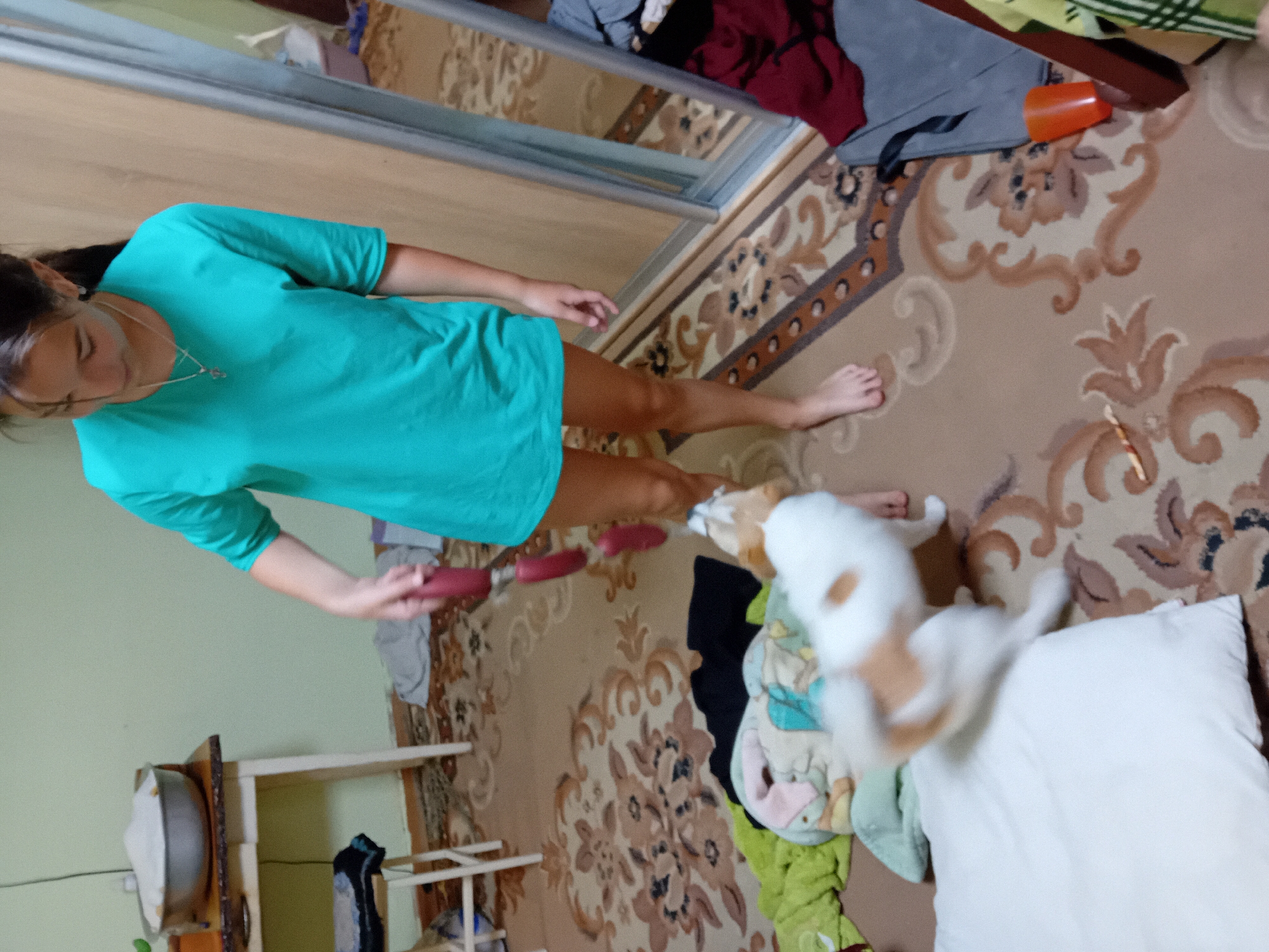 A young female is playing with a small dog in a room with a patterned carpet and light green walls. A wooden table with a large metal bowl filled with white powder is against the far wall. A stool with clothes on it is to the left of the table. In the foreground, a white and brown dog is on the floor, and a white pillow is nearby. The female is wearing a teal t-shirt and shorts. The dog is facing away from the camera, jumping. The environment appears to be a domestic interior. The location is identified as Chuhuiv, Ukraine.