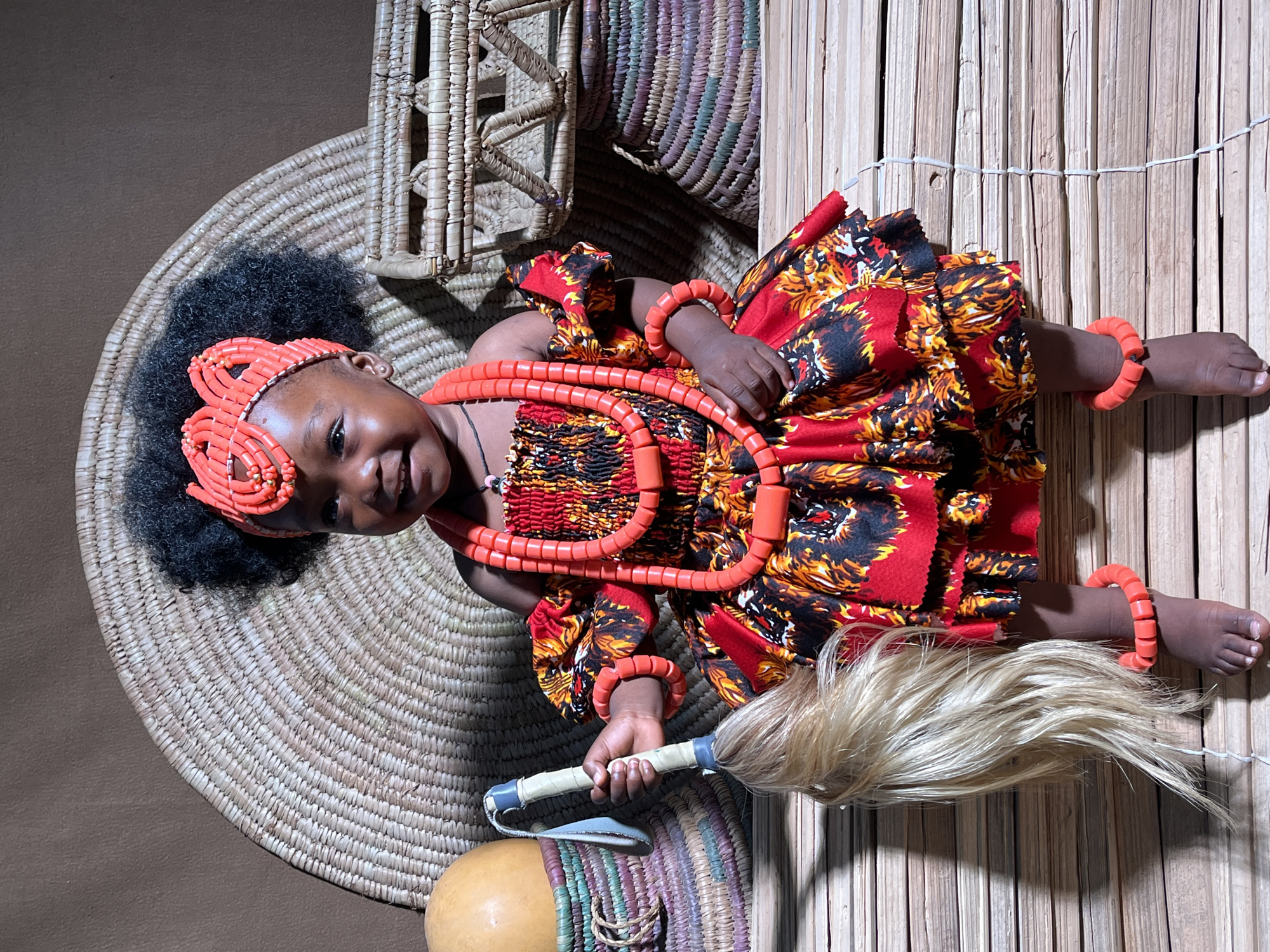 A young girl wearing traditional Nigerian clothing lies on a woven mat, holding a ceremonial staff. She is adorned with red beaded necklaces, bracelets, and a headband. She is positioned on a round mat and a bamboo structure is in the background. A calabash gourd is near her. The location is Unknown, Unknown.