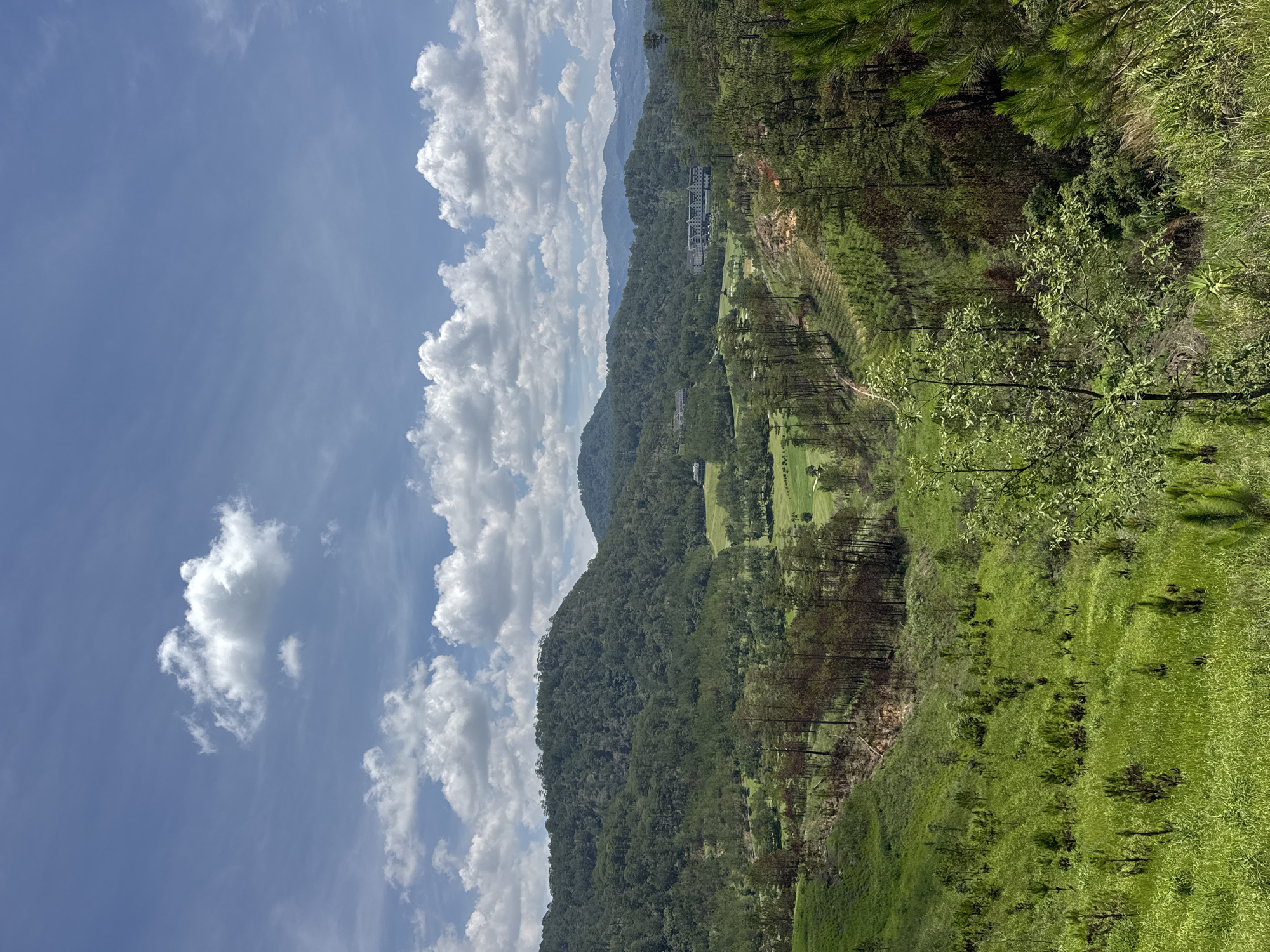 The image shows a landscape view in Hiep Thanh, Vietnam. In the foreground, there is green grassy terrain with scattered trees. Beyond that, a dense forest covers the lower slopes of a mountain. A golf course is visible with manicured greens and fairways amidst the trees. A multi-story building sits higher on the mountainside. The sky is blue with puffy white clouds.