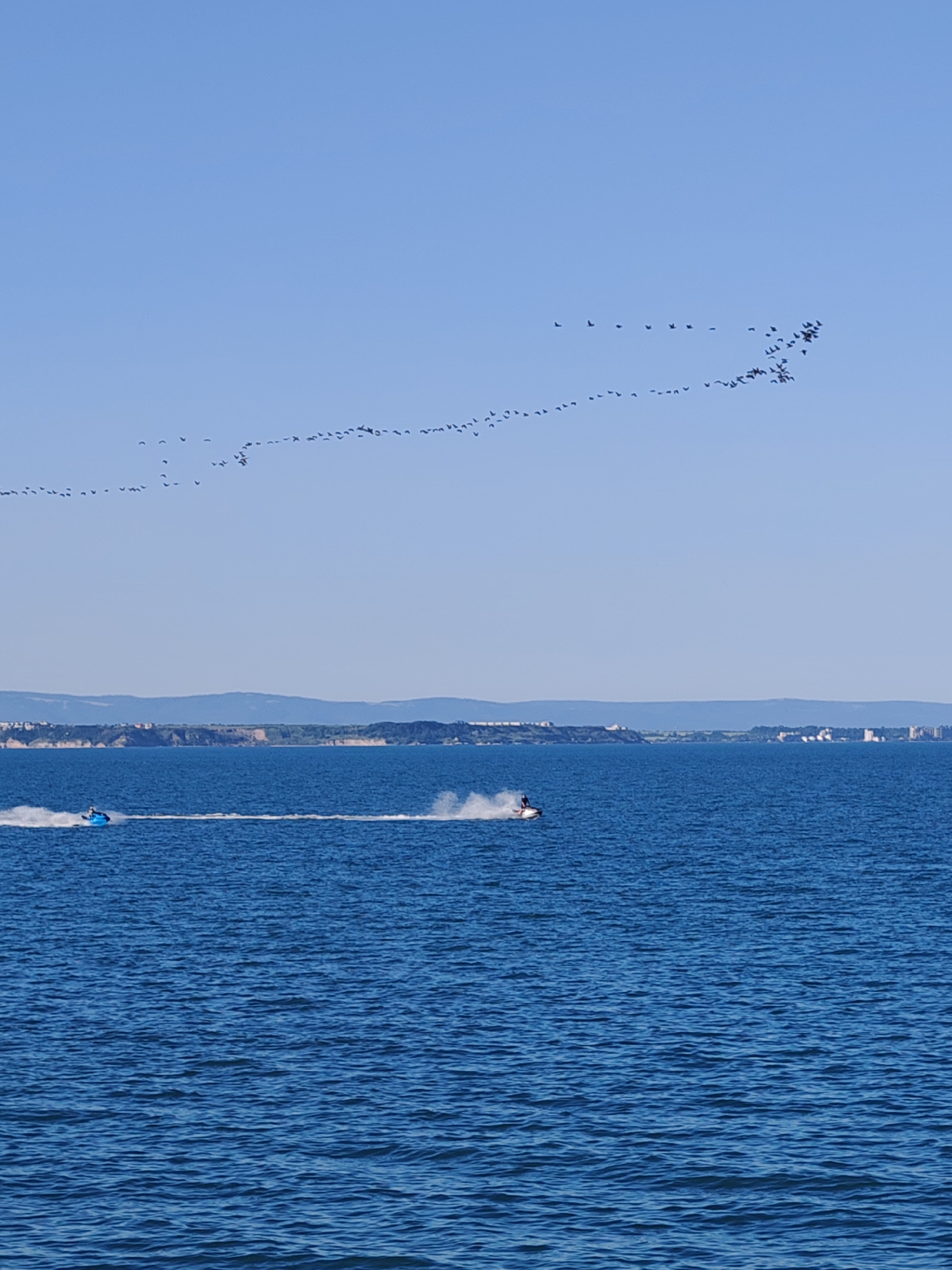 The image shows a blue sky with a long, sparse formation of birds flying across it. Below the sky, a body of water, possibly the Black Sea, stretches to a coastline with visible buildings and trees. Two jet skis are active on the water. One on the left is blue, pulling a blue inflatable tube, while another on the right is black with a single rider. The location is likely near Burgas, Bulgaria, given the landscape and architectural details visible on the coastline.