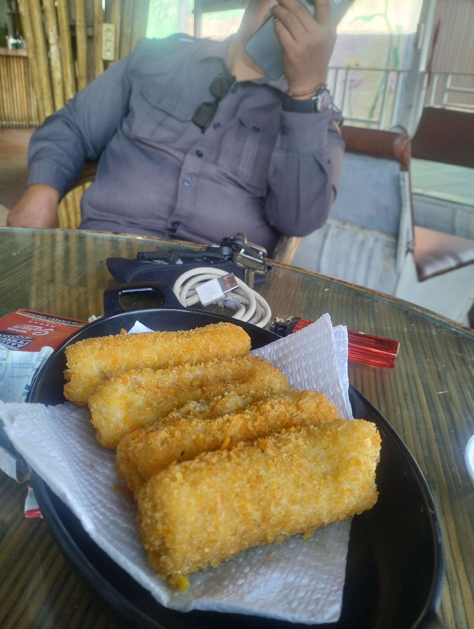 A person is seated at a table in what appears to be an outdoor cafe or restaurant with a bamboo wall in the background. The person, wearing a grey shirt, is holding a mobile phone to their ear. In the foreground, a black bowl contains several golden-brown, breaded snacks, possibly croquettes or spring rolls, resting on a white paper napkin. A red lighter and a white charging cable are visible on the table. The table has a translucent surface with a wood-grain pattern. The image is taken from a slightly elevated, close-up perspective, focusing on the food. The location is identified as Uteungeulinggang, Indonesia.