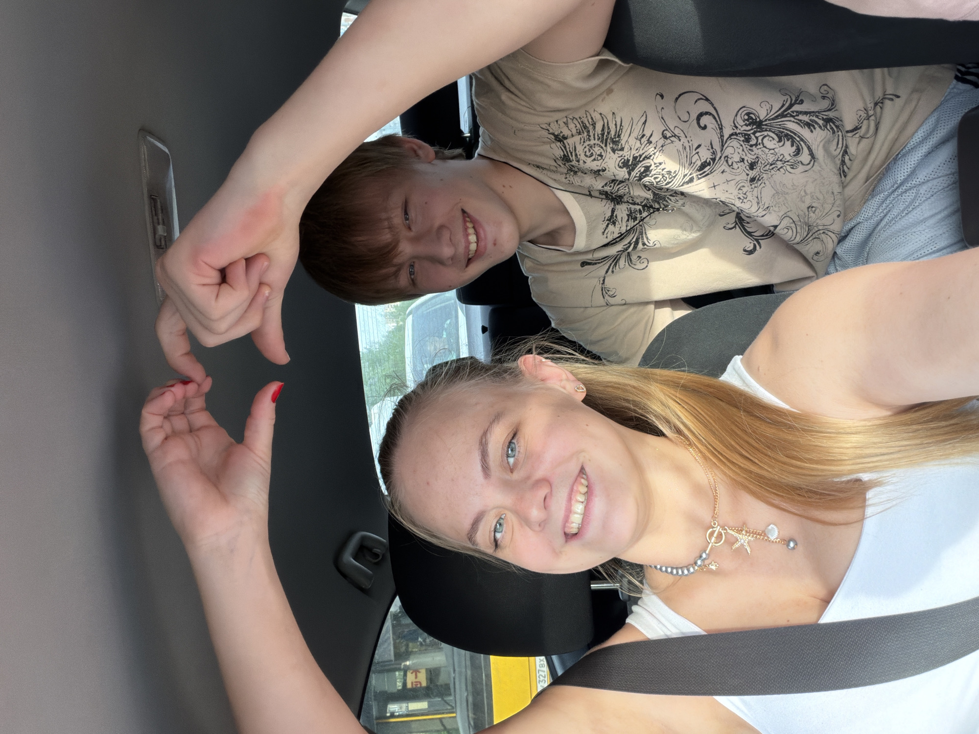 Two young people are posing for a selfie inside a vehicle. The female subject is in the driver’s seat wearing a white tank top and a necklace, secured by a seatbelt. She is holding up her left hand, with visible red nail polish. Her fair hair is blonde.
The male subject is behind her, wearing a beige t-shirt with a dark design. He is holding up his right hand. Together, their hands form a heart shape.
The vehicle’s interior is visible, with grey panels. Outside the window, a glimpse of a yellow vehicle with the license plate "327 EX" can be seen. The vehicle is presumably in Izhevsk, Russia, given the prompt's instructions.