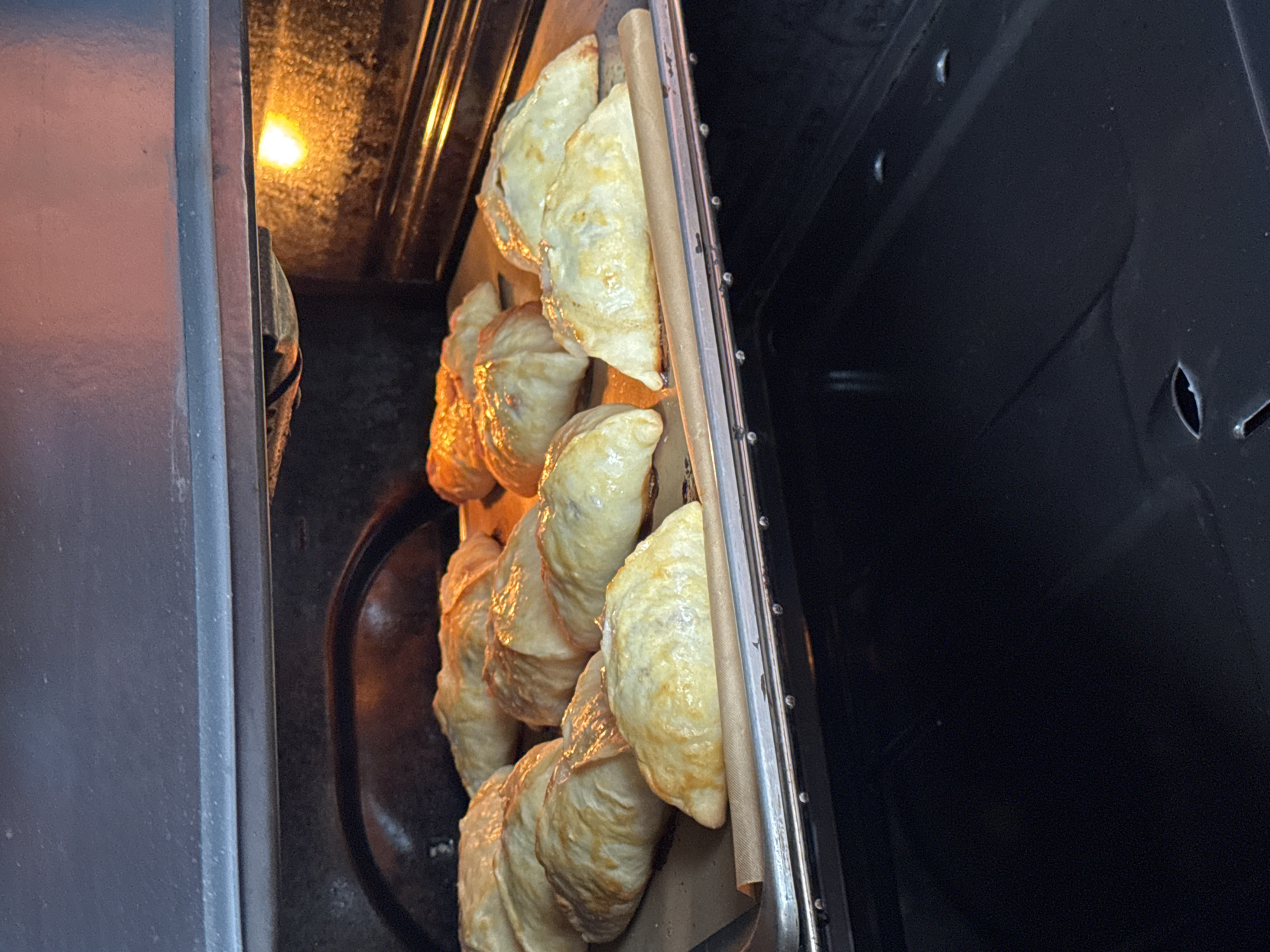 The image shows a close-up view inside an oven, presumably in Kaduy, Russia. Several crescent-shaped pastries, likely empanadas or pierogi, are arranged on a baking sheet lined with parchment paper. The pastries closest to the oven door are golden brown, while those further inside appear lighter in color. The oven's interior is dark, with a light bulb visible in the back. The oven door is slightly ajar.