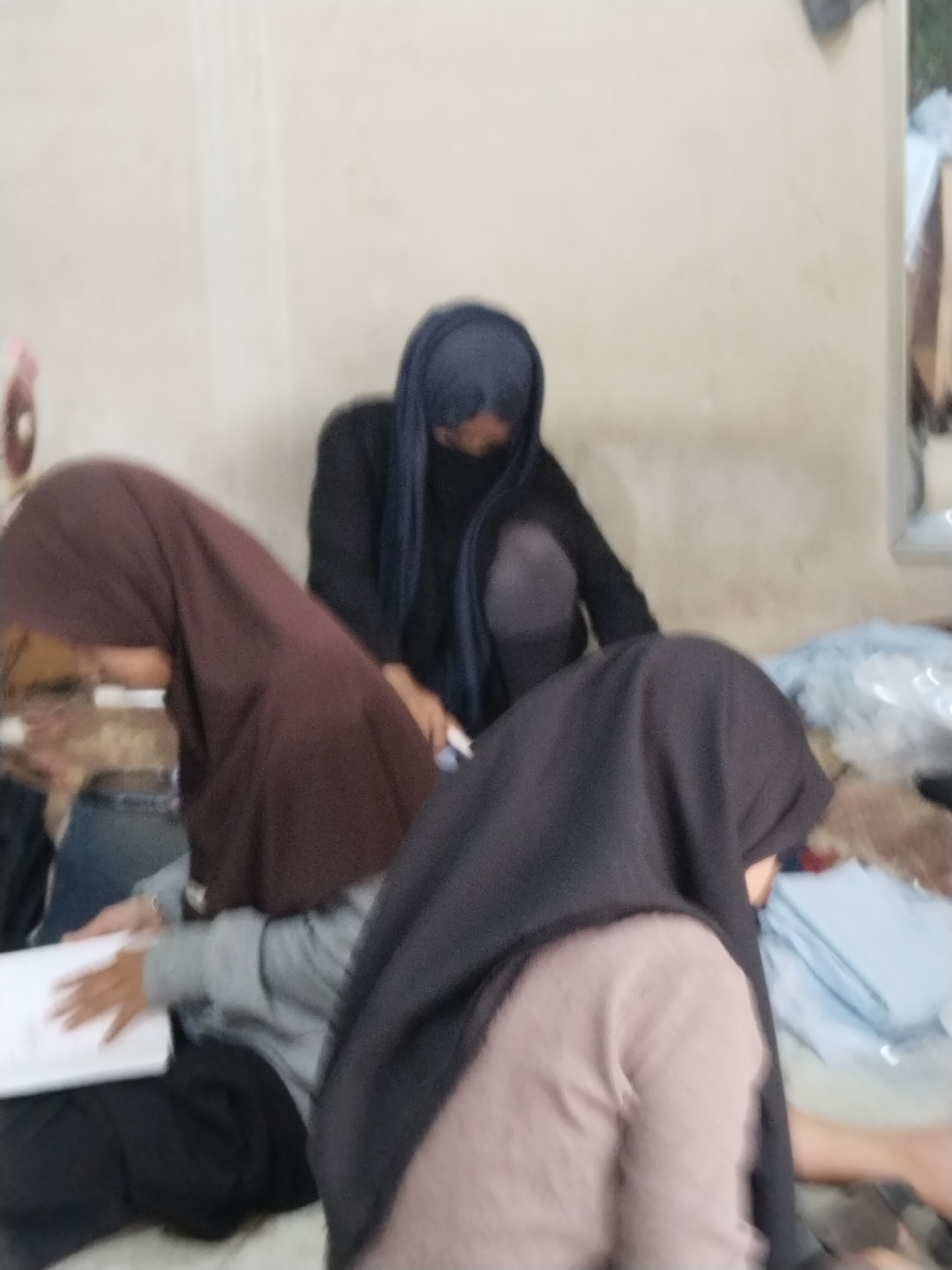 The image shows a blurry indoor scene with three people wearing hijabs. One person in the foreground on the right wears a black hijab and a light pink top. Another person in the center wears a blue hijab and a black top. The third person on the left wears a maroon hijab and a grey top. They are seated, and one person on the left is holding a white notebook. The environment appears to be a room with a light-colored wall and some stacked fabric items in the background. The image is low resolution and lacks clear details. The location is listed as Unknown, Indonesia.