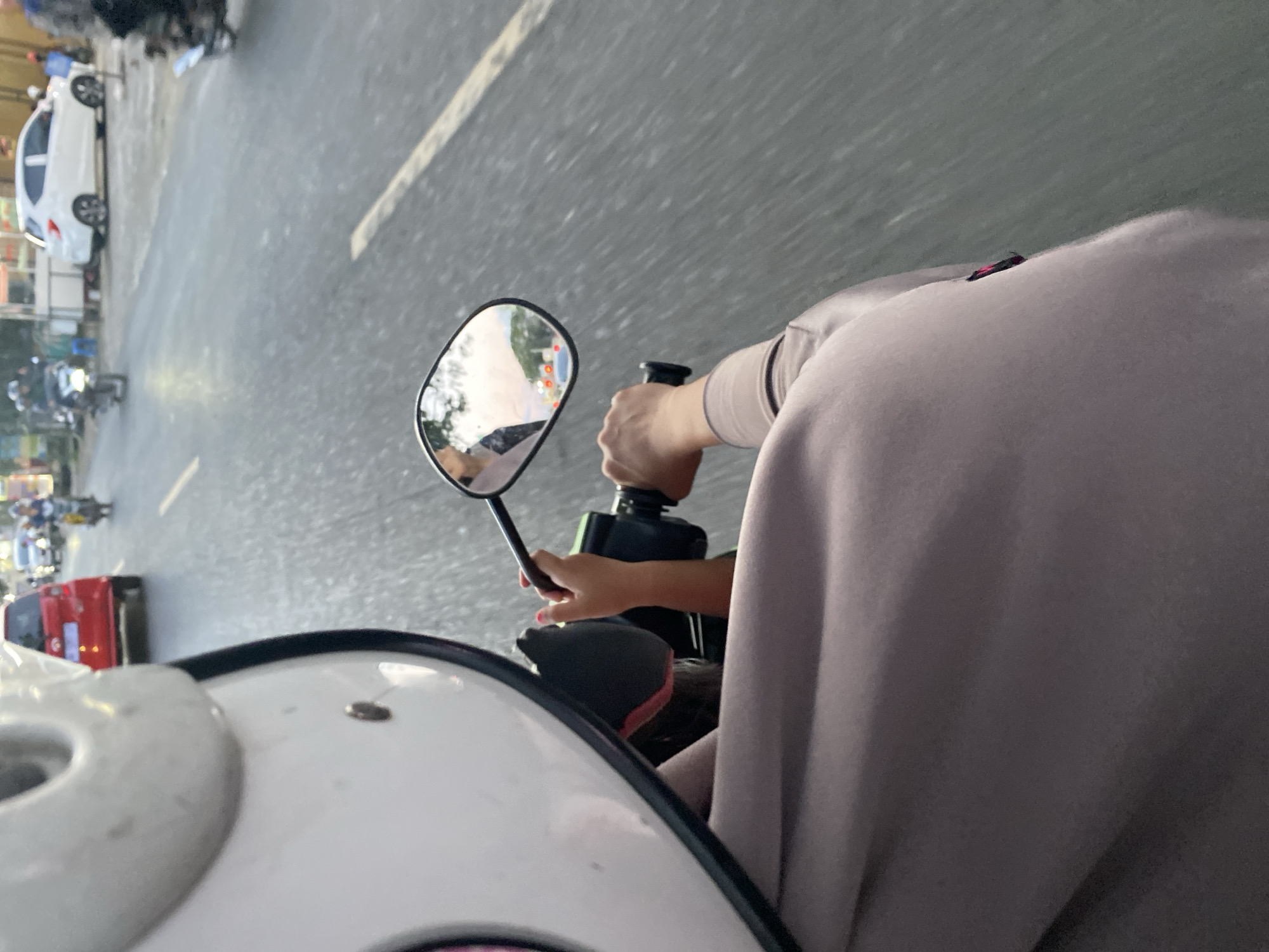 A POV shot captures a person riding a motorcycle on a street in Macege, Indonesia. The rider, wearing a white helmet and a light brown hoodie, is holding the handlebars. A child's small hand, with painted nails, is also visible reaching towards the handlebars. In the rearview mirror, a reflection shows a person in the back and vehicles on the road, including a red car and other motorcycles. The environment appears to be a moderately busy street with trees and buildings lining the roadside. The sky suggests it may be late afternoon or early evening.