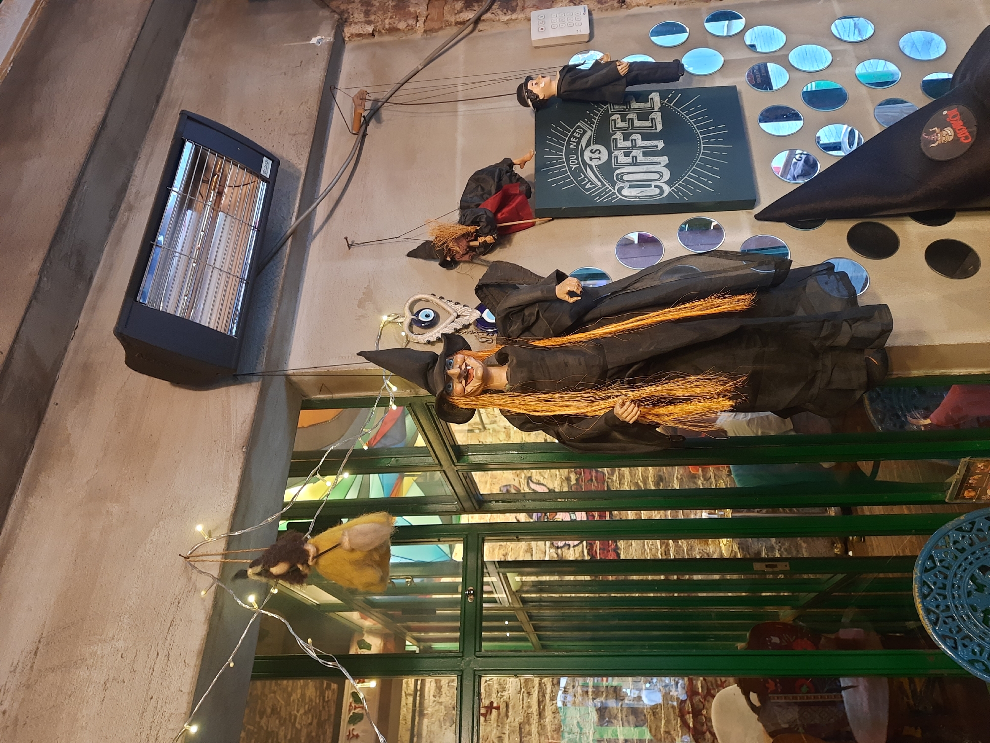The image depicts the interior of a cafe in Kadıköy, Turkey, decorated for Halloween. A green metal-framed structure with glass panels spans the scene. Mounted on a wall are several Halloween decorations: a black electric heater, a doll resembling a man in a suit, a witch on a broom, and a large witch figure with long orange hair. A sign with the word "COFFEE" is also visible. String lights are strung near a yellow bird decoration. Below, through the glass panels, are glimpses of interior decor, including tables and chairs.