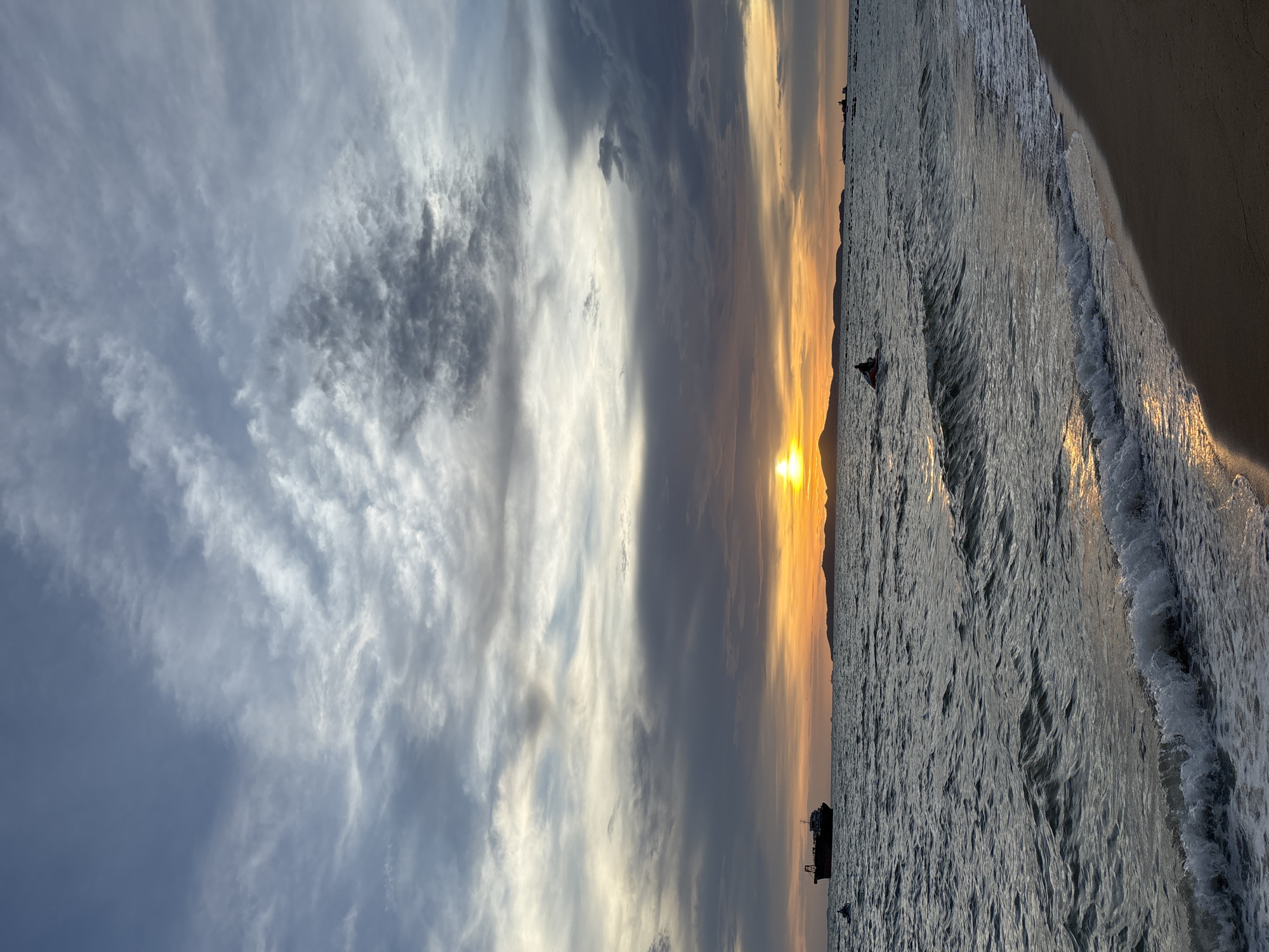 The image depicts a sunset scene at Pattaya Beach, Thailand. The sun is low on the horizon, casting a warm orange glow across the sky and water. Dark, wispy clouds streak across the sky, creating a dramatic backdrop. In the foreground, waves gently lap against a sandy shore. Further out at sea, a large cargo ship is visible on the left, and a person is riding a jet ski in the middle distance. Several other boats and distant landmasses can be seen on the horizon. The overall atmosphere is serene and picturesque.