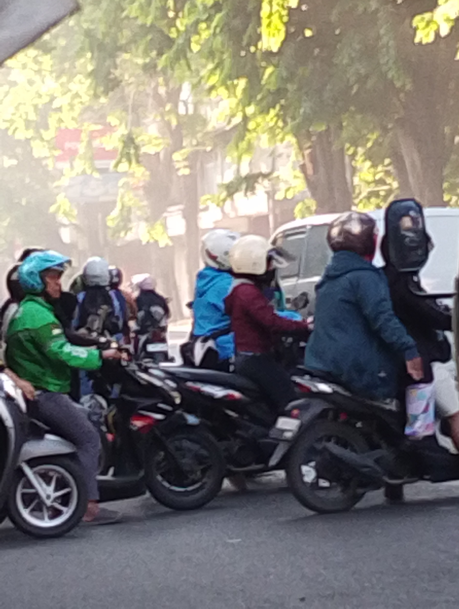 The image depicts a street scene in Bangil, Indonesia, where several individuals are riding motorcycles. The foreground is dominated by two motorcycles with riders. The motorcycle on the left has a rider wearing a green jacket and a blue helmet. The motorcycle on the right has a rider in a dark blue jacket and a brown helmet, with another person behind them wearing a black helmet and mask. In the background, more motorcycles and riders are visible, as well as a white van. Trees with green foliage line the street, and sunlight filters through the leaves, creating a dappled effect. The road surface is asphalt. The scene appears to be a typical daytime street activity in a Southeast Asian city.