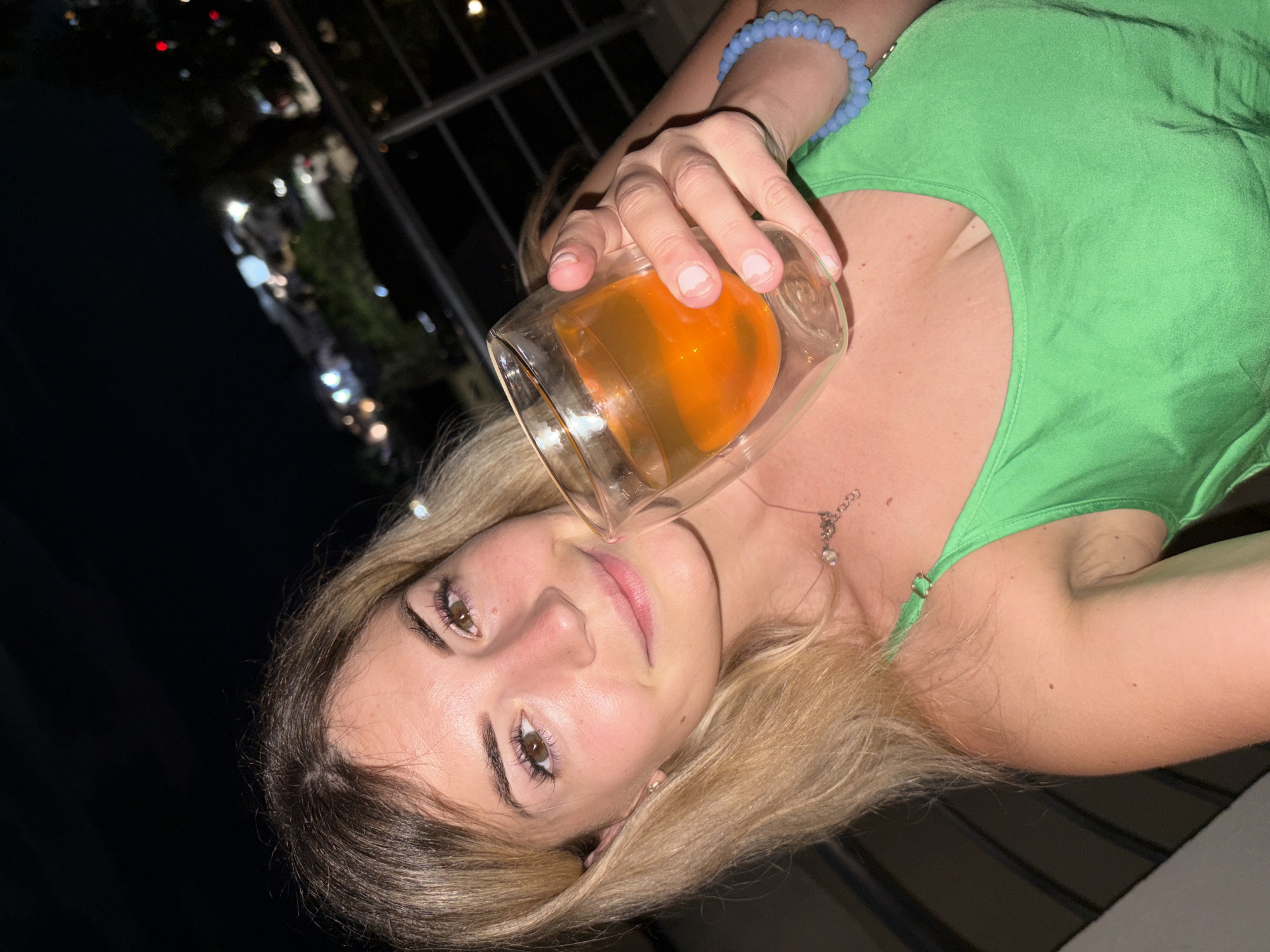 The image features a young woman with blonde hair holding a glass of an orange beverage. She is wearing a green tank top and a blue beaded bracelet. The scene appears to be outdoors at night, with blurred lights in the background suggesting a city or town setting. The woman is looking towards the camera with a slight smile, and her expression conveys a sense of relaxation or enjoyment. The lighting is dim, characteristic of nighttime, and there are no visible signs of specific weather conditions. The overall mood is casual and celebratory. There is no visible text in the image.