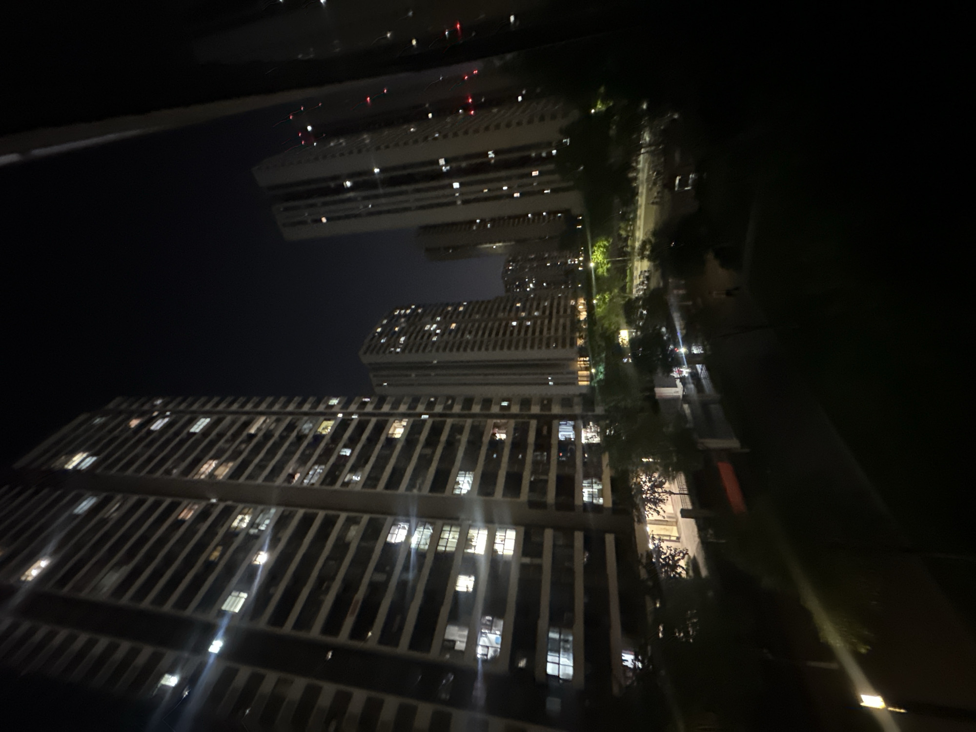 This is a nighttime photograph depicting a dense urban landscape in Chongqing High-tech Zone, China, likely captured from an elevated position. The image is oriented such that multiple tall buildings stand vertically against a dark, clear night sky.
In the foreground, a large high-rise building occupies the lower portion of the frame. Its facade is characterized by numerous rectangular windows, many of which are brightly lit, indicating occupancy and activity within. The building exhibits a modern architectural style with prominent vertical and horizontal structural elements, possibly balconies or decorative fins. Light flares and reflections are visible around some of the illuminated windows, adding to the nighttime glow.
Further in the midground, several other high-rise structures are visible, receding into the distance. These buildings also display numerous glowing windows, suggesting a bustling residential or commercial area. Red lights, possibly aircraft warning lights, can be seen atop one of the more distant tall buildings, piercing the darkness.
Running through the middle distance, a street or thoroughfare is illuminated by streetlights, casting a warm, yellowish glow on the pavement and surrounding trees. The trees line parts of the street, and faint blurs of light might indicate moving vehicles, though no distinct cars or people are discernible. The sky above is a deep, dark blue-black, devoid of clouds.
The overall scene conveys a sense of a vibrant, active metropolis at night, with the myriad of lights from buildings and streets creating a striking visual contrast against the dark environment. There is no visible text in the image.
