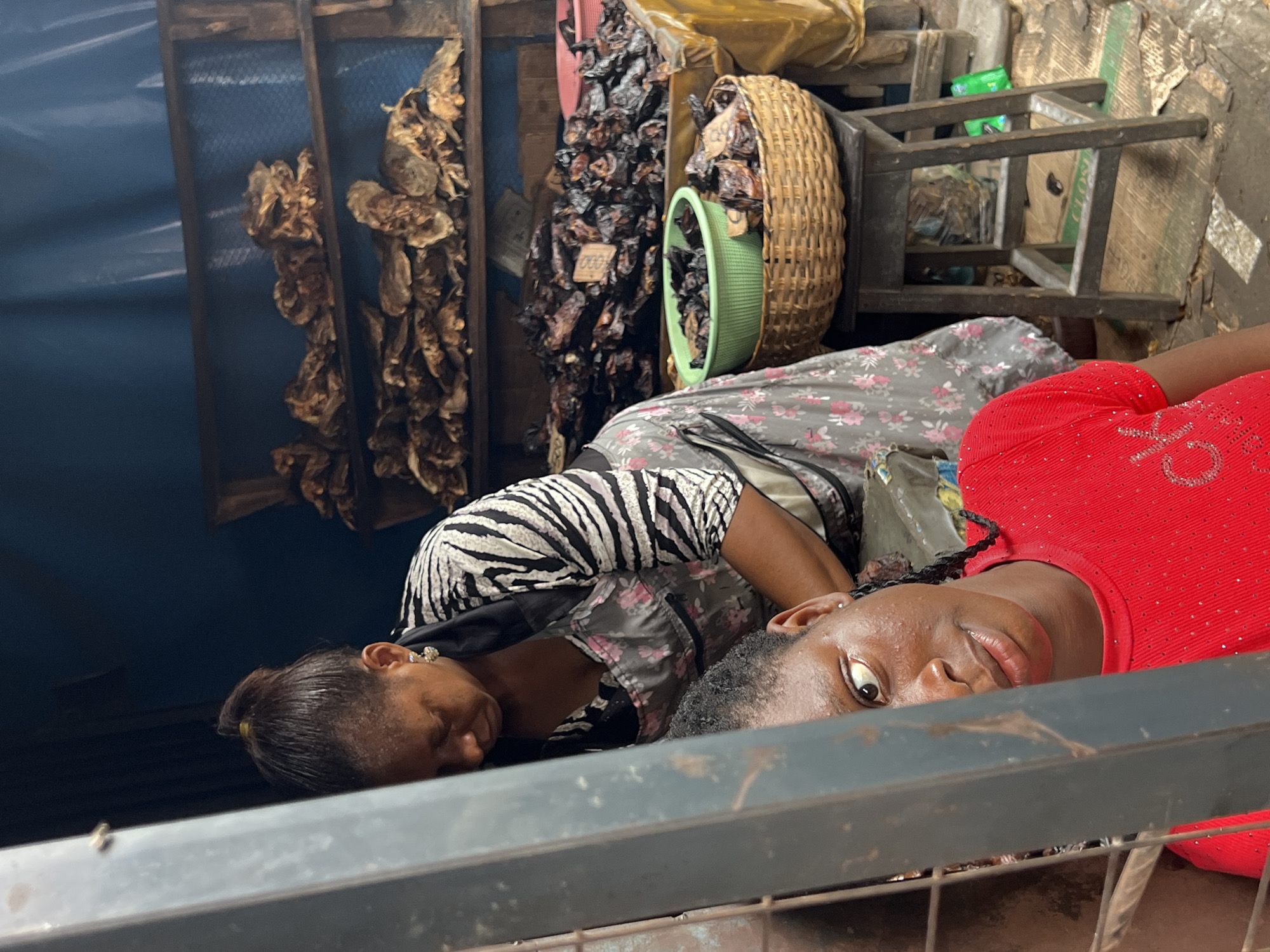 In Nkpor, Nigeria, a market scene unfolds as two women are captured in a close-up shot. The woman in the foreground, wearing a vibrant red shirt adorned with tiny shimmering embellishments, is partially obscured by a dark metal structure. Her gaze is directed towards the viewer, with one eye prominently visible, conveying a direct and perhaps curious expression. Her braided hair is visible.
Behind her, another woman, dressed in an apron with a floral pattern over a black and white striped top, is engaged in an activity. Her head is bowed as she works, her facial features largely in shadow. She wears a shiny earring.
The background reveals a stall laden with dried fish, stacked on wooden shelves. Another basket, overflowing with similar dried fish, sits on a wooden stool, alongside a green plastic bowl. The environment appears to be an outdoor market setting, suggested by the rustic wooden structures and the assortment of goods. The overall lighting is somewhat dim, contributing to the candid nature of the photograph.