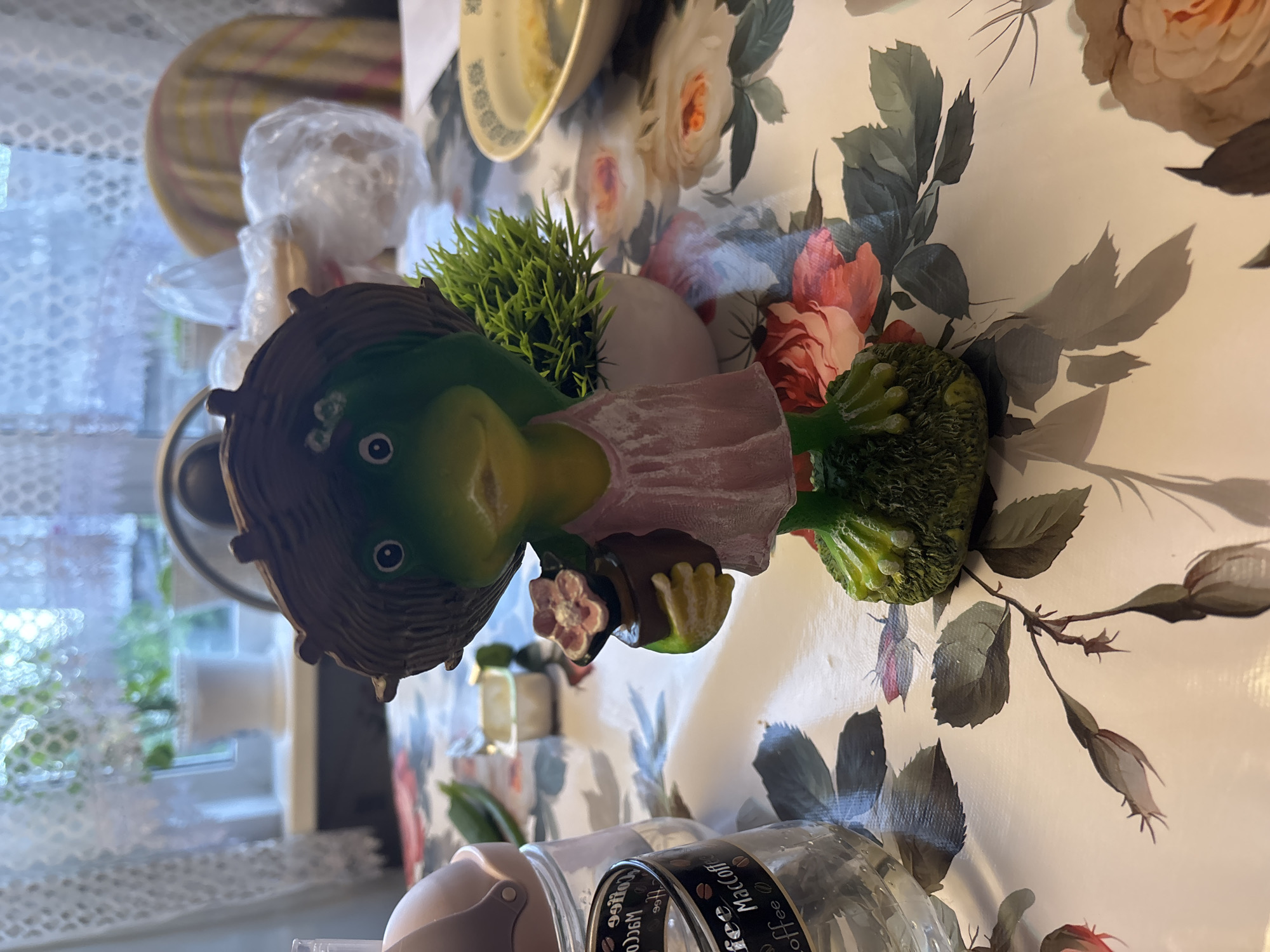 The image shows a figurine of a frog wearing a straw hat and a pink dress, holding a small potted flower. The figurine is placed on a table covered with a floral-patterned tablecloth. In the background, there is a window with lace curtains, a potted plant, and some plastic-wrapped items. There are also a glass jar with coffee branding and a bowl with some food on the table. The scene appears to be indoors, possibly in a residential setting. There is no identifiable location in the city of Kurgan, Russia.