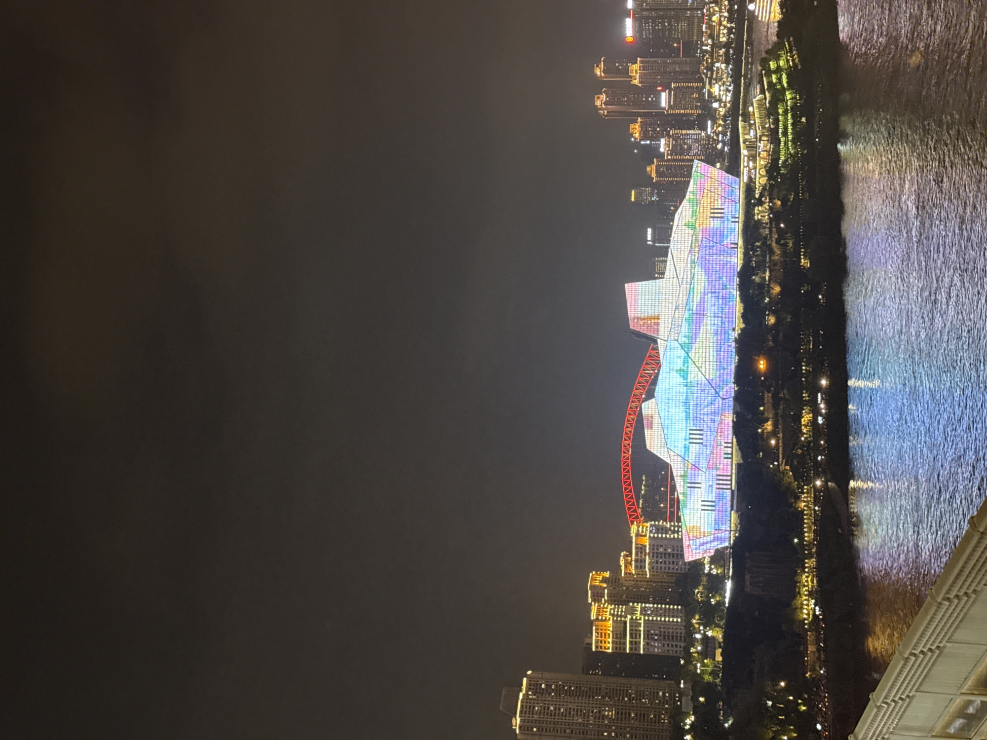 The image depicts a nighttime scene in Yuzhong District, China, showcasing a vibrant cityscape along a waterfront. The central focus is a large, uniquely shaped building illuminated with a colorful, geometric light display, resembling a faceted gem or a crystalline structure. Above this building, a bright red, arched bridge spans across the frame. To the left of the illuminated building are several tall, modern skyscrapers, some with visible windows lit from within, and others adorned with decorative yellow and white lights. To the right of the central building, more high-rise structures rise into the dark sky, also illuminated. A body of water, reflecting the city lights with streaks of blue and purple, occupies the foreground. The shoreline is lined with trees and smaller illuminated structures, suggesting a park or landscaped area. The overall impression is one of a bustling, modern urban environment at night, with a strong emphasis on architectural lighting and a scenic waterfront.