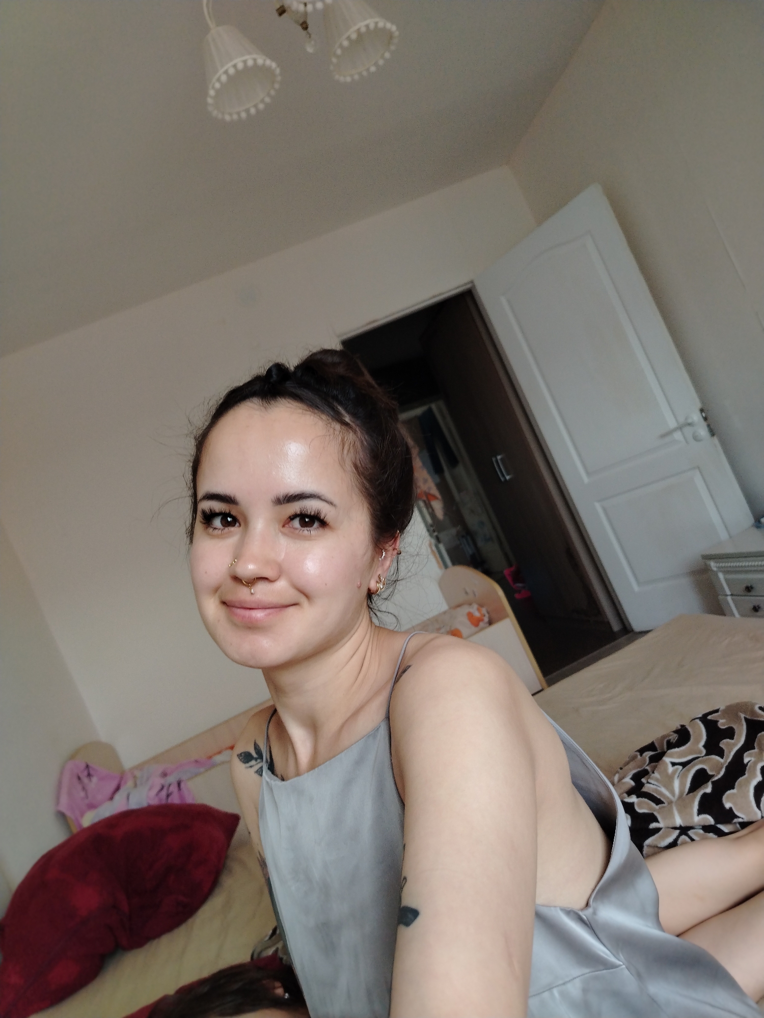 A young woman with dark hair pulled into a bun and a nose ring is seen from the chest up, smiling into the camera. She is wearing a light gray satin camisole and has a tattoo on her left arm. The background suggests an indoor domestic setting, possibly a bedroom, with a bed, pillows, and a closet visible. The lighting appears natural, coming from a window or overhead source. The image does not provide information about the specific location within Noviy, Russia.