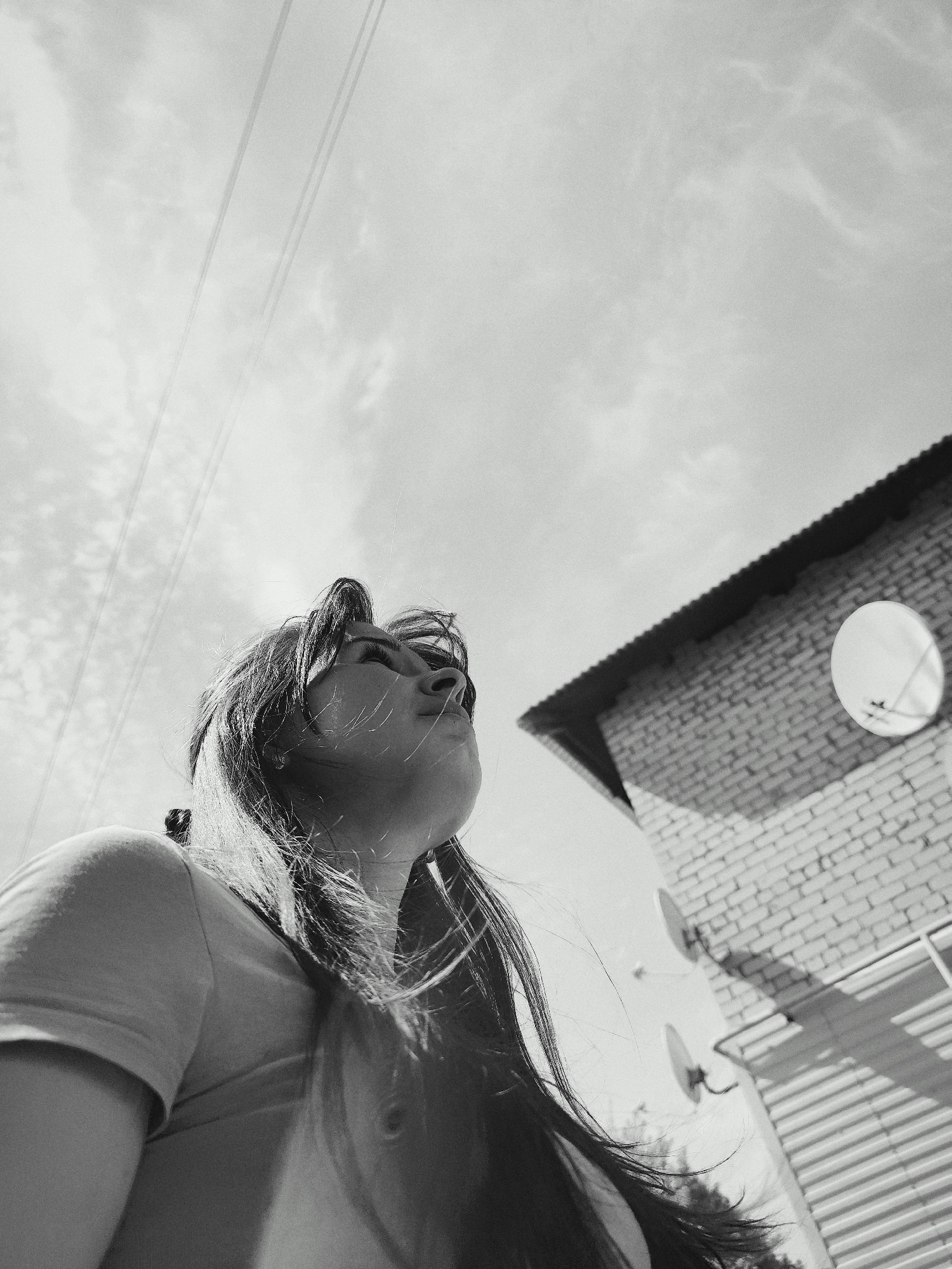 The black and white image presents a low-angle view of a woman looking up towards a sky filled with clouds, with some utility lines visible overhead. The woman has long hair that is partially obscuring her face. To the right of the frame, there is a brick building with a satellite dish attached to the wall. Another satellite dish is visible lower on the wall. There is a corrugated metal surface visible below the building. Based on the reference to Фоки, Russia, it can be inferred that this photograph was taken there.