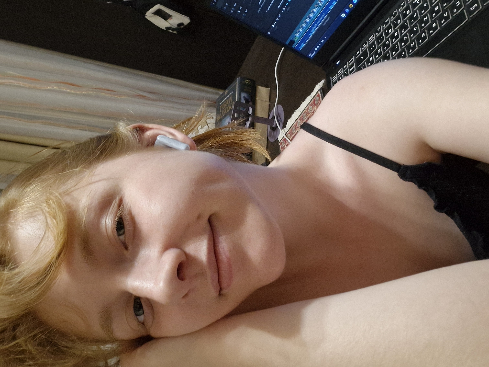 A pale-skinned person with fair hair lies on a bed or soft surface. They are wearing a black top with spaghetti straps. An earbud is visible in their ear. To the right, a black laptop computer rests on a dark surface. Behind the laptop, there are books and a small device. A partially visible patterned surface is in the background. It is not possible to discern the location as Yekaterinburg, Russia from the image.