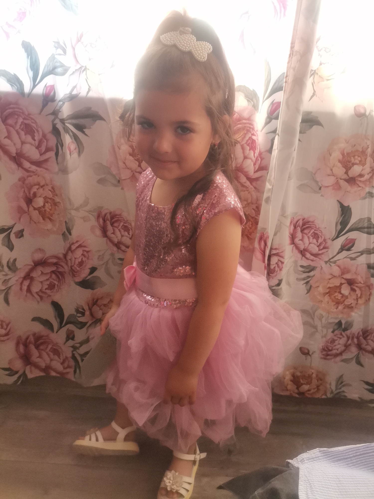 A young girl stands indoors in front of a floral print backdrop. She is wearing a pink dress with a sequined bodice and a layered tulle skirt, paired with white sandals. Her hair is partially up with a beaded hair accessory. The setting, location, and city are unknown.