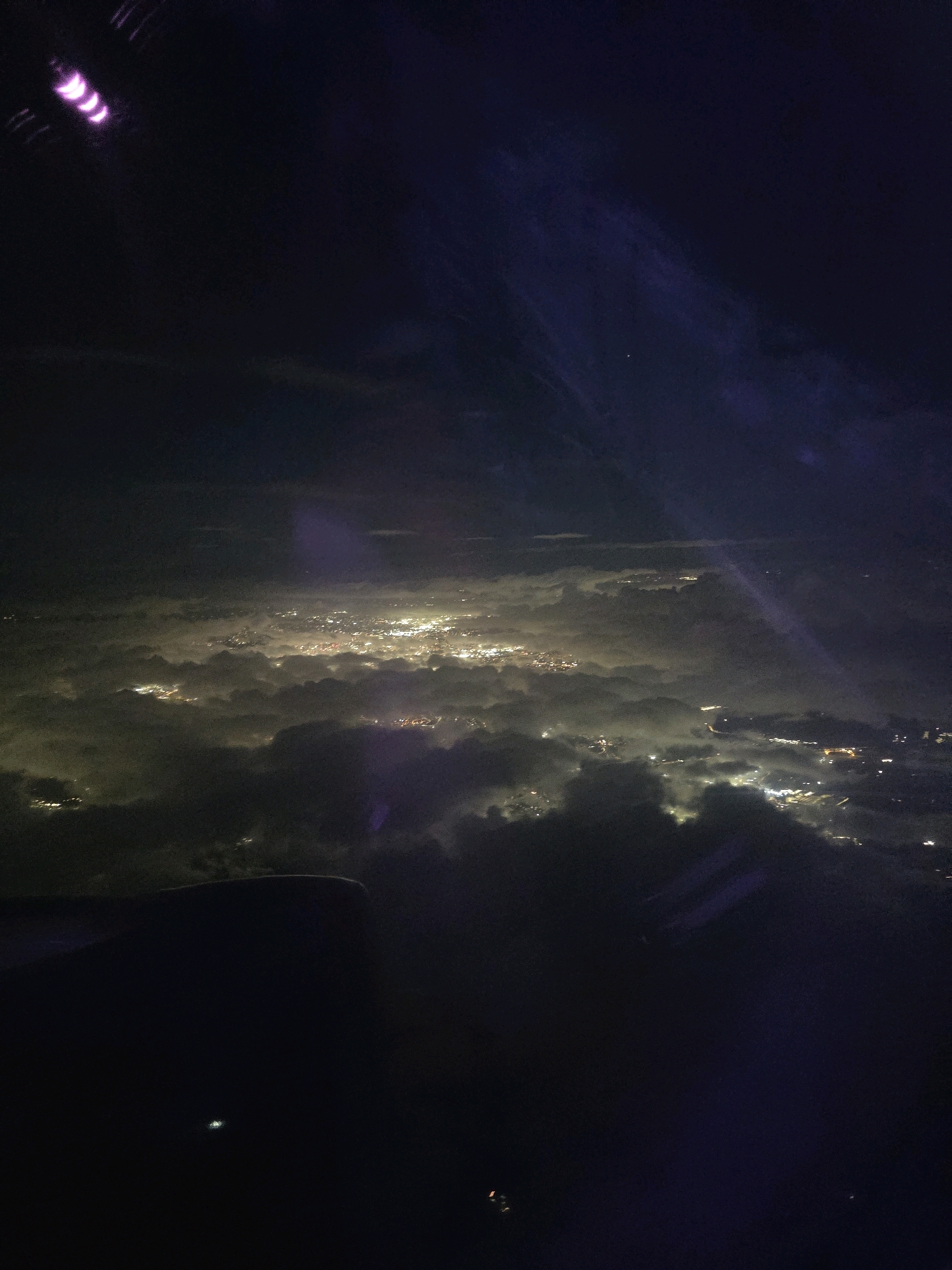 An aerial view from an airplane window at night showcases a sprawling cityscape illuminated by countless lights, peeking through a thick layer of clouds. The lights form clusters and streaks, suggesting the presence of roads and buildings, indicating a densely populated urban area. The clouds, illuminated from below by the city lights, appear as a soft, glowing blanket, with darker pockets of shadow. The upper portion of the image is dark, with a faint purple hue and a streaky light phenomenon, possibly a reflection or atmospheric effect. A portion of the airplane's wing or fuselage is visible in the lower left corner, silhouetted against the darker background. In the far upper left corner, a bright purple, crescent-shaped light with a swirling effect is visible. The lights from the city are concentrated in areas, suggesting neighborhoods, commercial districts, and industrial zones. There is no visible activity of people or any specific objects that can be identified, other than the implied urban infrastructure. The scene is one of a vast, unseen city shrouded in darkness and clouds, with its lights offering a glimpse of human presence. This appears to be a view over a large urban area, possibly within the wider region of North Warwickshire, United Kingdom, given the context.