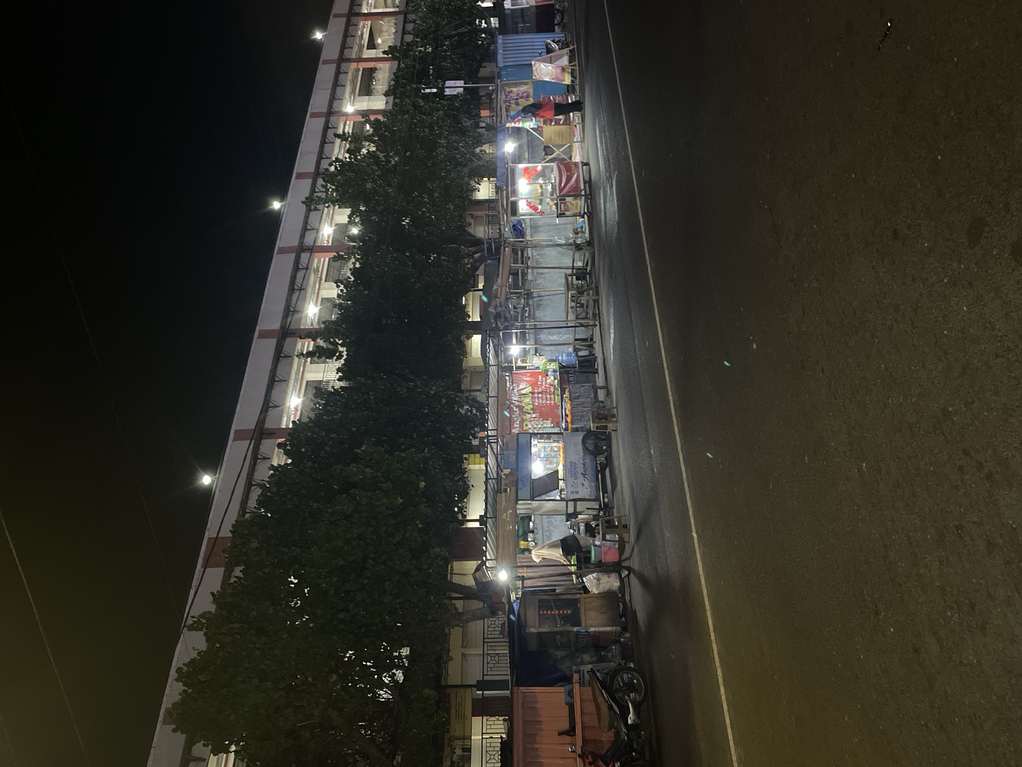 The image shows a street scene at night in an urban setting. In the foreground, a dark asphalt road is visible, with a white line marking the lane. Behind the road, a row of food stalls or small shops line the sidewalk. These stalls are illuminated by artificial lights and have various signs and displays. Several people are present, either tending to the stalls or interacting with customers. To the left, a motorcycle is parked. In the background, a large, multi-story building stands, with lights visible from within and along its exterior. Trees are planted in front of the building, their dark foliage contrasting with the illuminated structures. The overall atmosphere is that of a bustling night market or commercial area.