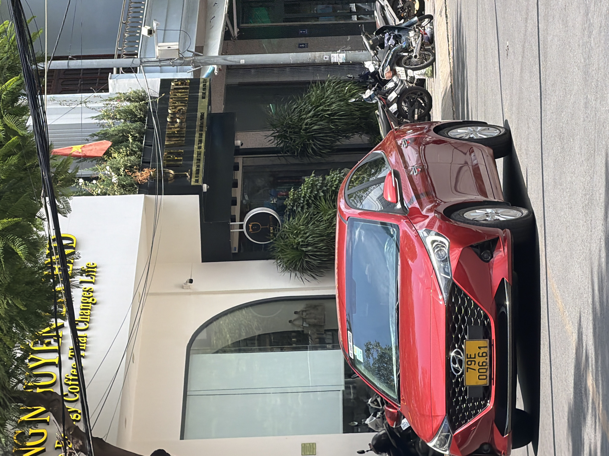 A red Hyundai sedan is parked on a street in Nha Trang, Vietnam, with a yellow license plate reading "79E 006.61". The car is positioned in front of a building with a white facade and large glass windows. Above the windows, signage in yellow lettering is visible. To the right of the building, a Vietnamese flag is displayed. Several motorcycles are parked on the side of the road. The environment suggests a commercial area with shops and businesses.