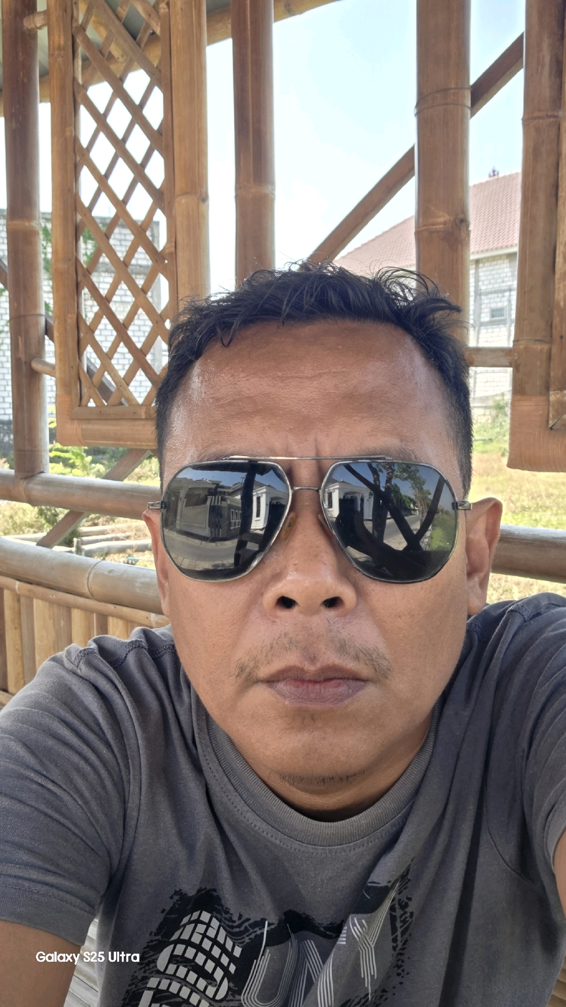 This is a selfie taken by an adult male with medium-toned skin, wearing a grey t-shirt and aviator sunglasses. He has short, dark hair and a mustache. He appears to be outdoors, seated in a structure made of bamboo, possibly a gazebo or veranda. In the reflection of his sunglasses, houses and trees are visible. The background also shows bamboo poles and a lattice-like structure, suggesting a rustic or natural setting. The lower left corner of the image displays the text "Galaxy S25 Ultra", indicating the device used to capture the photograph. The location is identified as Juwana, Indonesia.