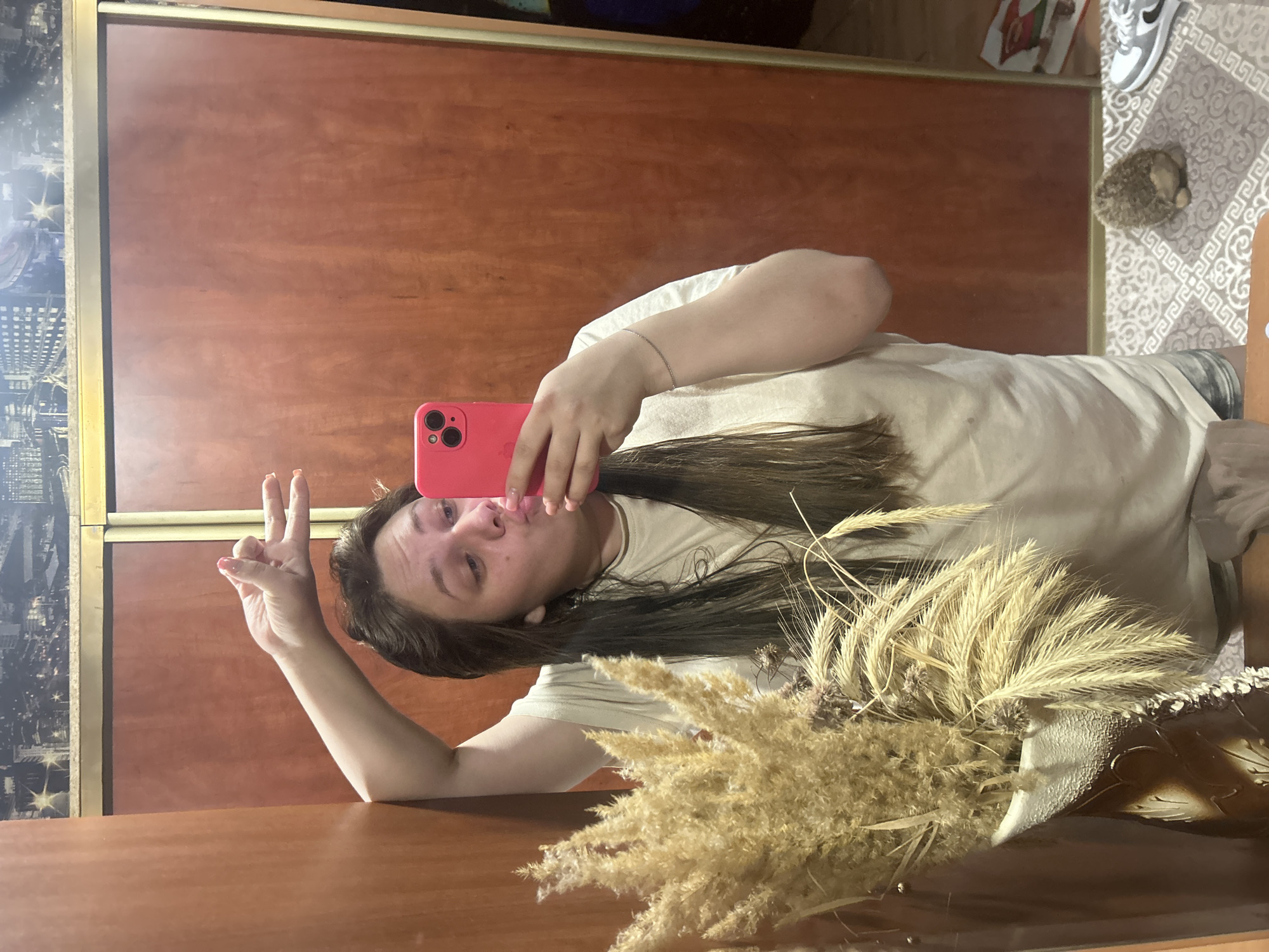 A person is taking a selfie with a pink phone in front of a mirrored closet. The person is making a kissing face and holding up a peace sign with one hand. A vase of dried wheat stalks is in the foreground. The floor has a rug with a geometric pattern, and there are shoes and a small round object on it. The closet has brown doors. There is a scene of a city visible on one of the closet doors. This image appears to be taken in an interior setting in a private dwelling. The location cannot be determined to be in Ольховка, Russia.