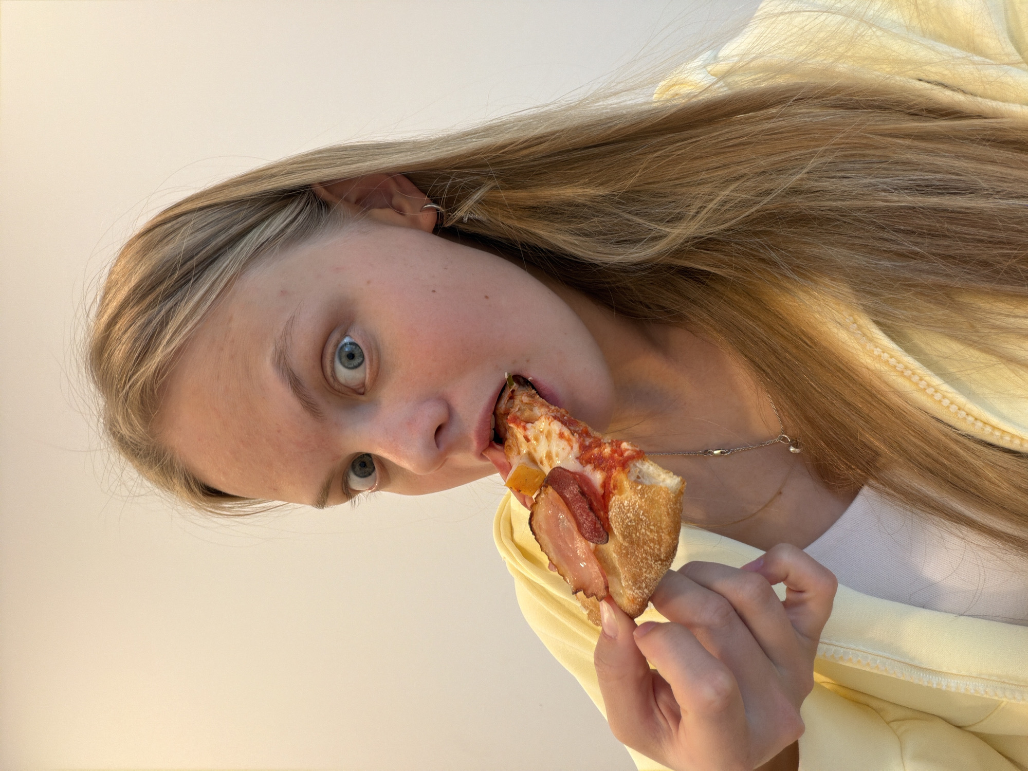 A young woman with blonde hair and blue eyes is eating a slice of pizza. The pizza has tomato sauce, cheese, pepperoni, bacon and bell peppers on it. She is wearing a yellow jacket. She also is wearing a necklace with a charm. The background is a plain white color. The location is unknown but is not Izhevsk, Russia.